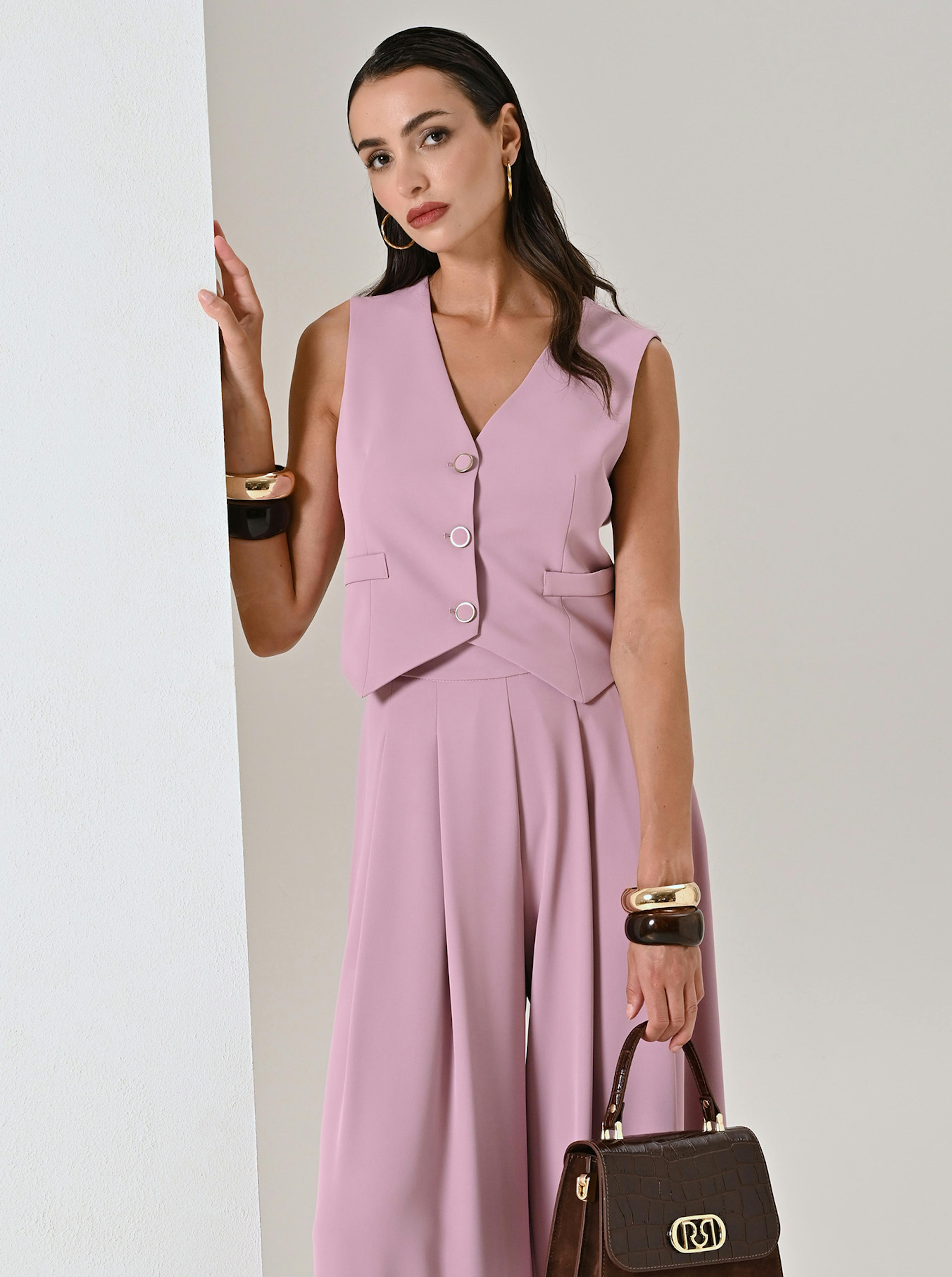 A woman in a stylish pink sleeveless suit with wide-legged pants leans against a white wall.