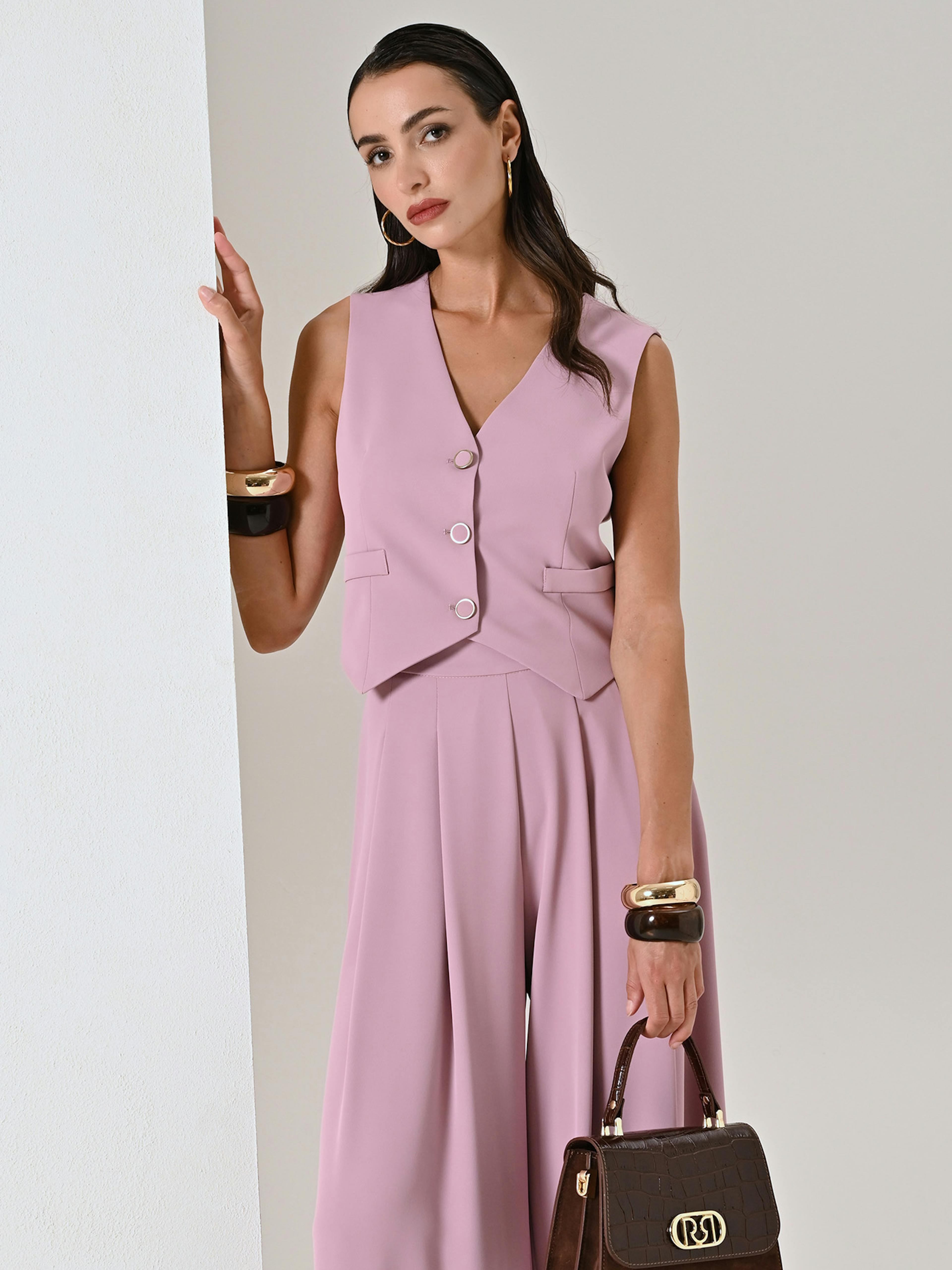 A woman in a stylish pink sleeveless suit with wide-legged pants leans against a white wall.