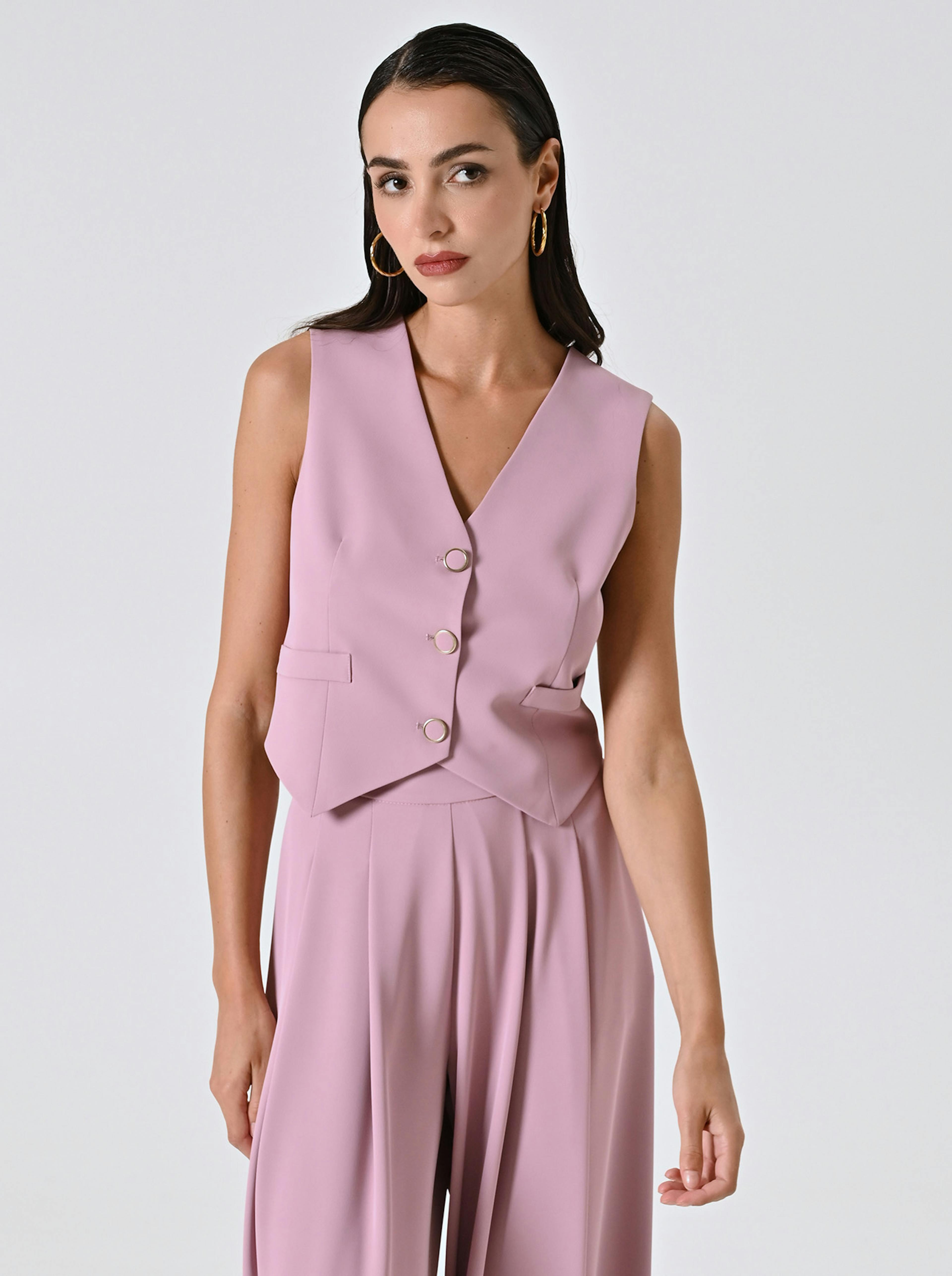 A person with long dark hair is wearing a sleeveless, light pink suit with a V-neck and gold buttons.