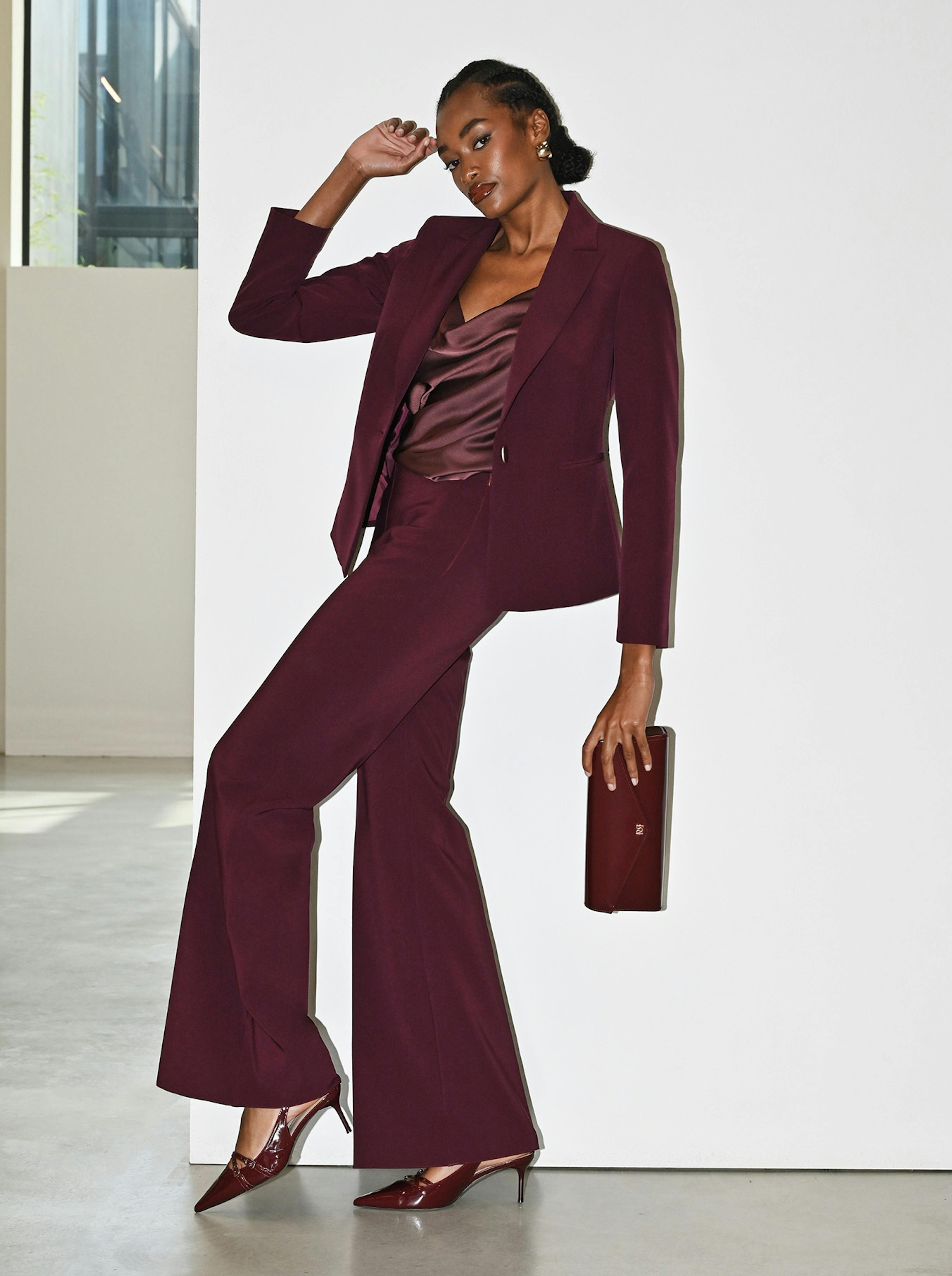A woman in a burgundy pantsuit leans against a white wall, holding a matching clutch. 