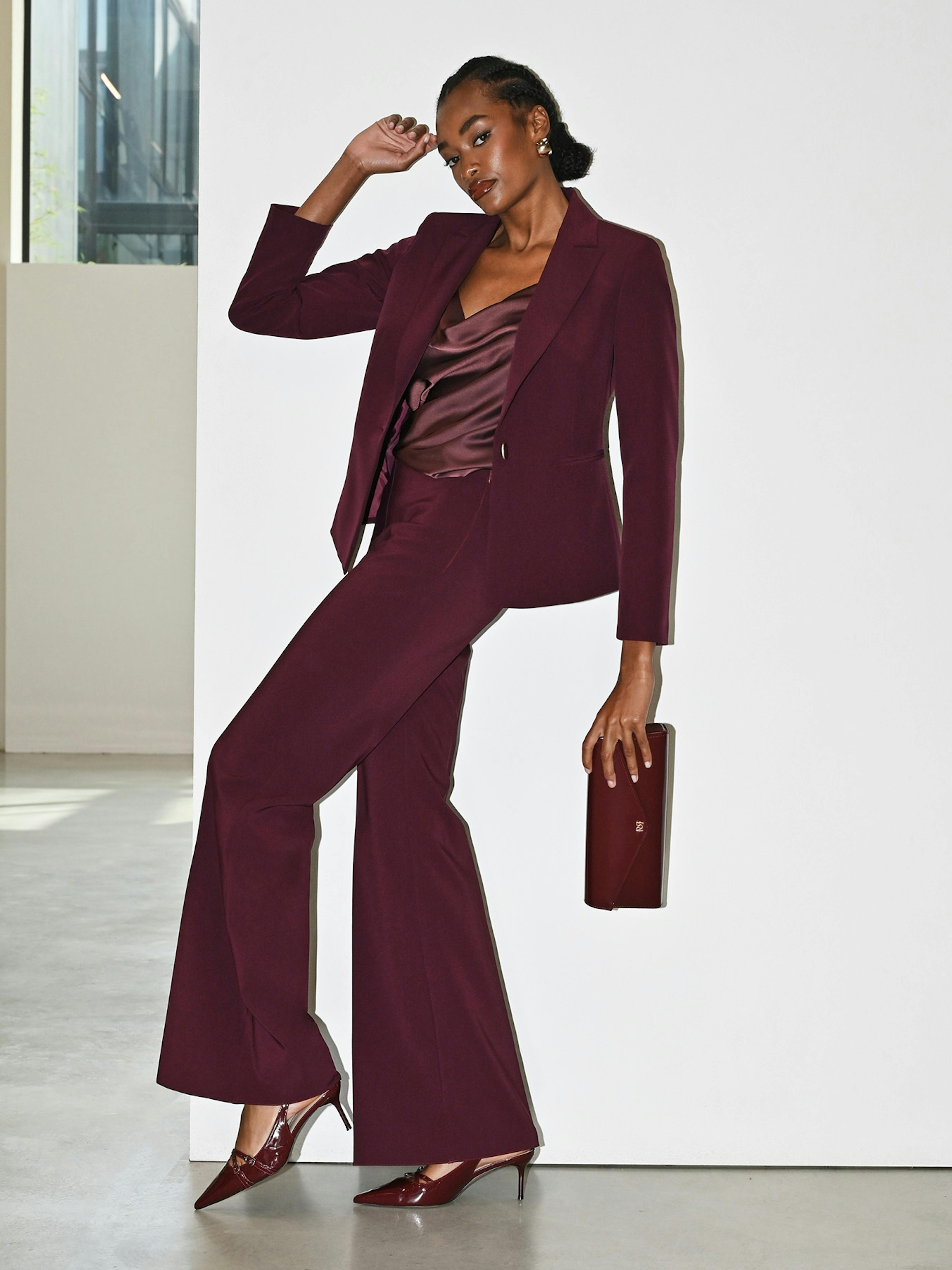 A woman in a burgundy pantsuit leans against a white wall, holding a matching clutch.