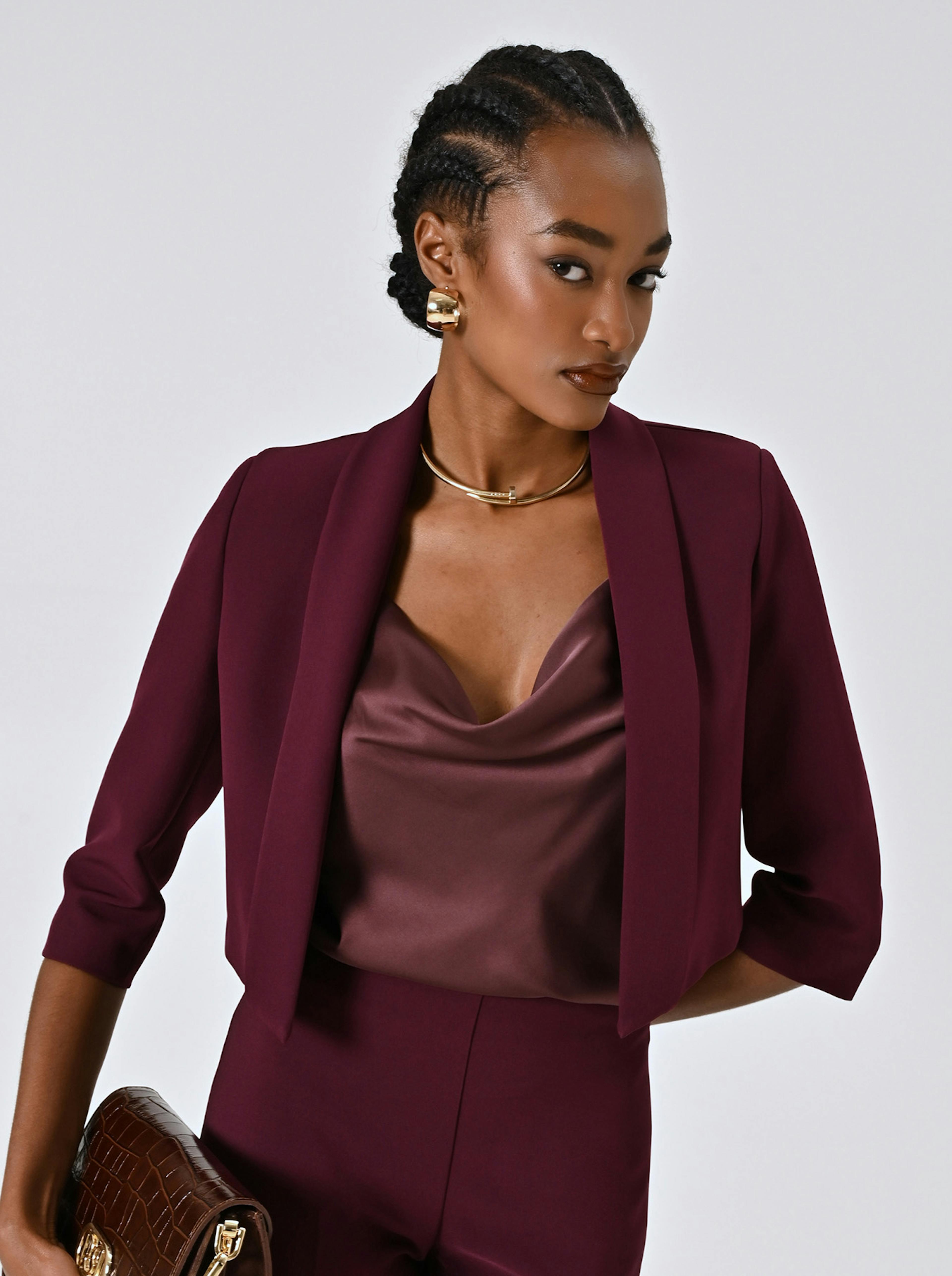 A confident person in a burgundy suit and silk top, accessorized with gold jewelry, holds a textured brown clutch against a neutral background.