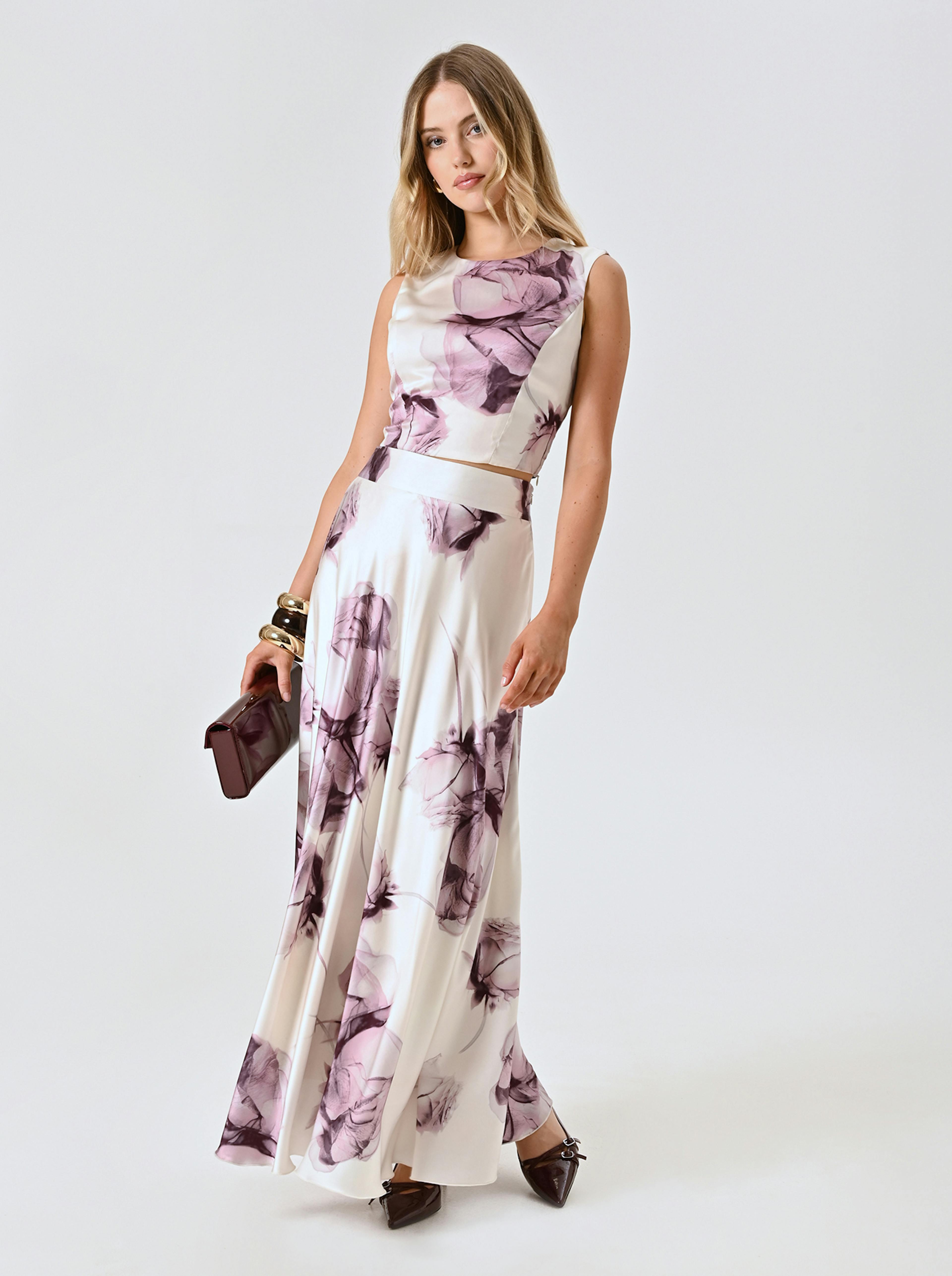 A woman poses in a sleeveless, white top and skirt with purple floral patterns. She holds a burgundy clutch and wears chunky bracelets, exuding elegance and poise.