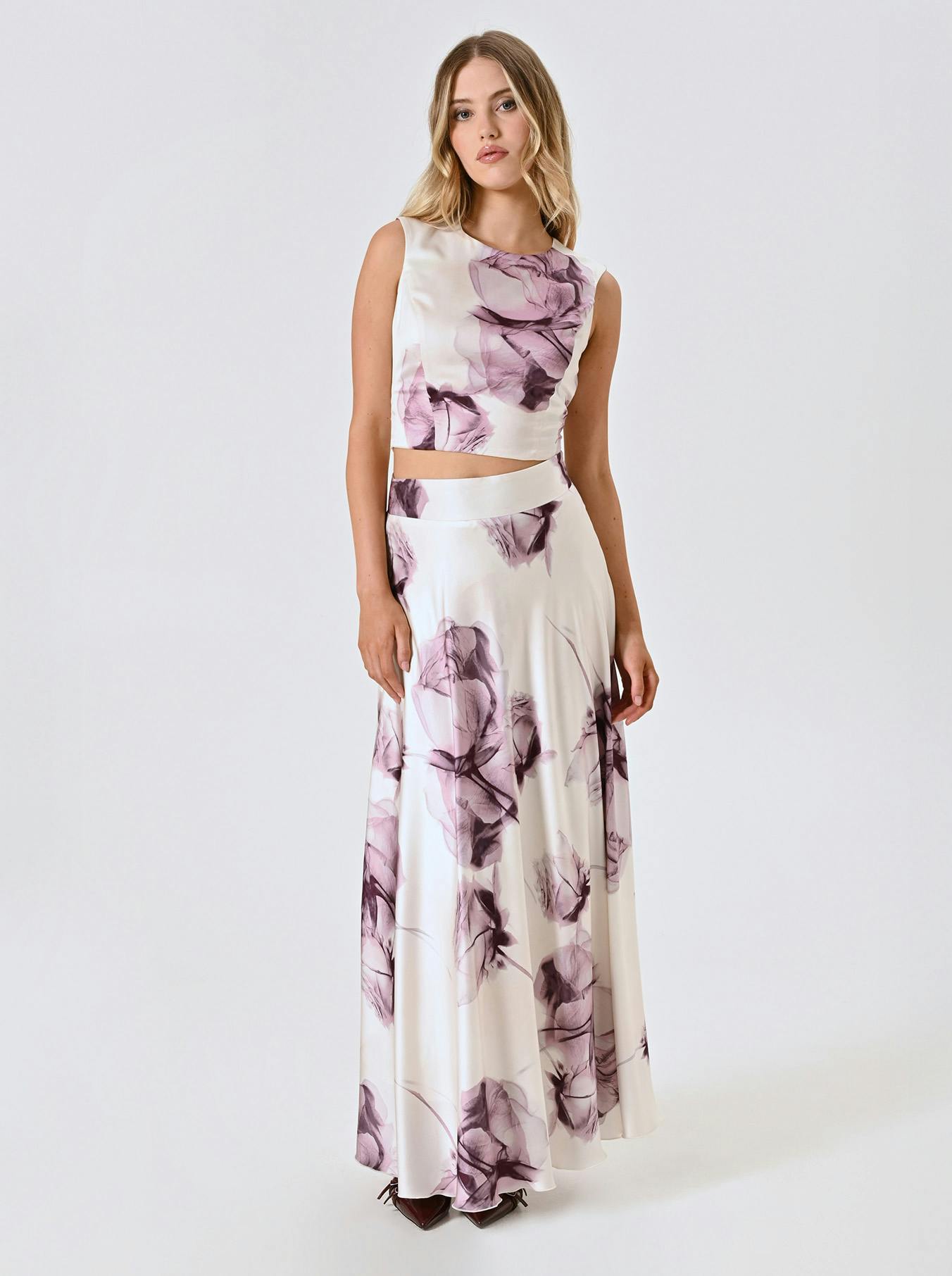 A woman poses in a sleeveless, white top and skirt with purple floral patterns.