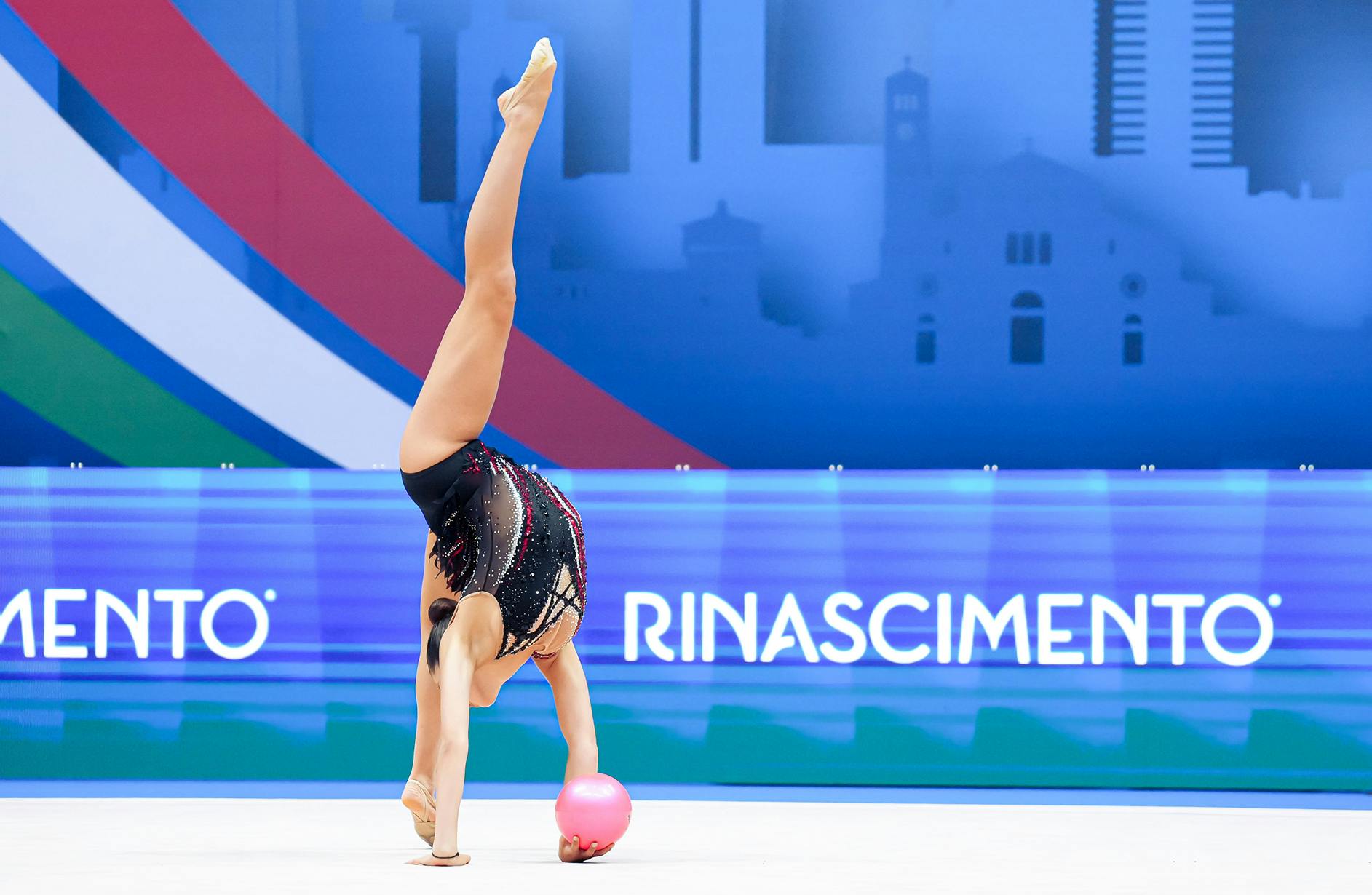 A rhythmic gymnast performs a one-handed handstand, balancing a pink ball, wearing a black, sparkling leotard. Background features city skyline art.