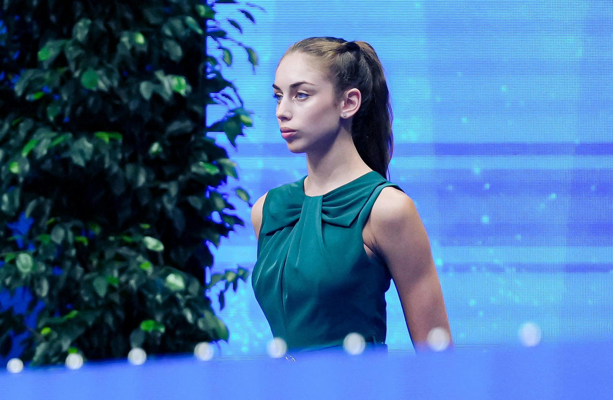 Young woman in a teal dress, standing against a vibrant blue background, appears focused and composed.