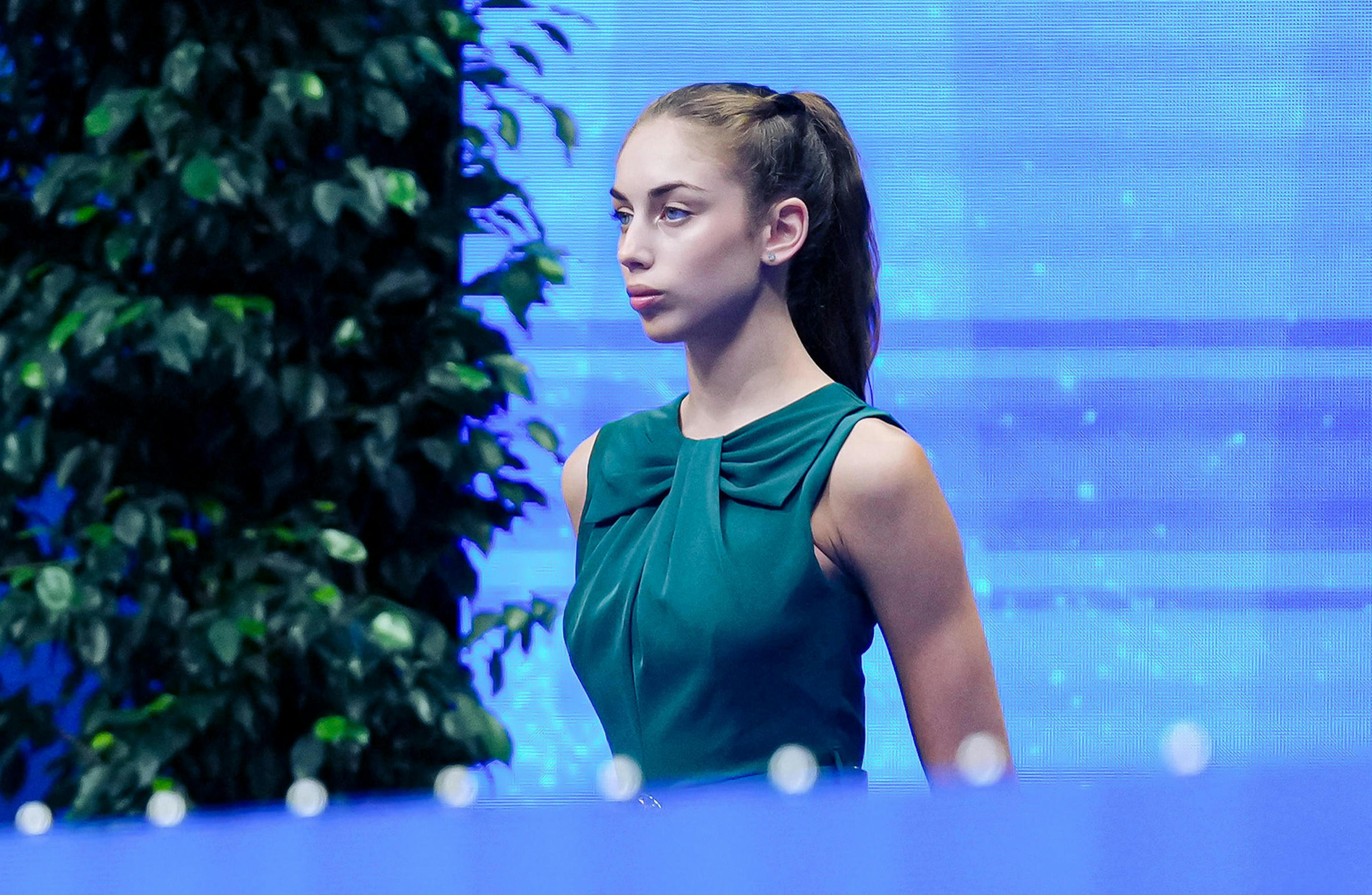 Young woman in a teal dress, standing against a vibrant blue background, appears focused and composed. 