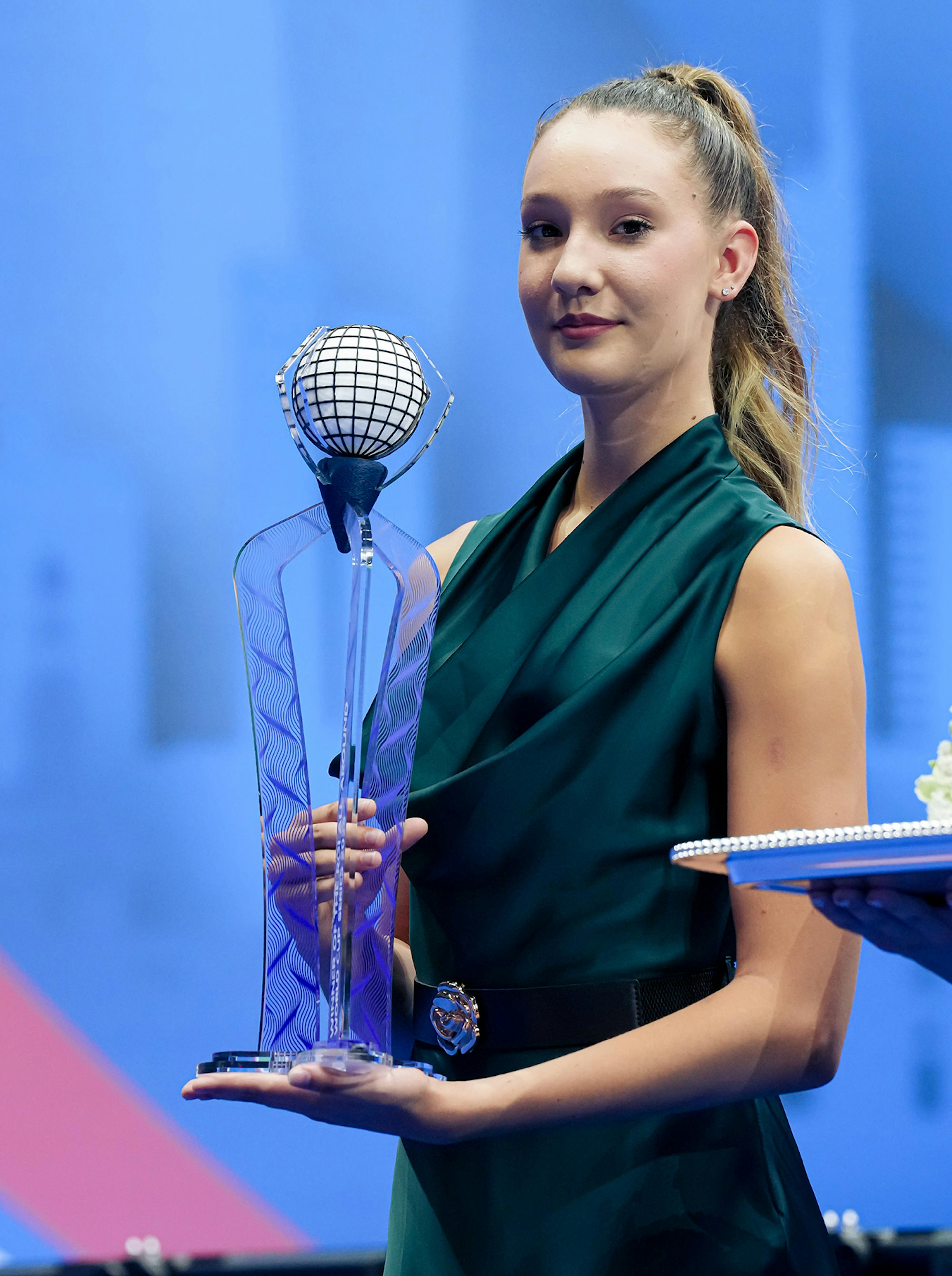 Young woman in a teal dress holds a tall, elegant glass trophy with a globe design. The background is a calm blue. 