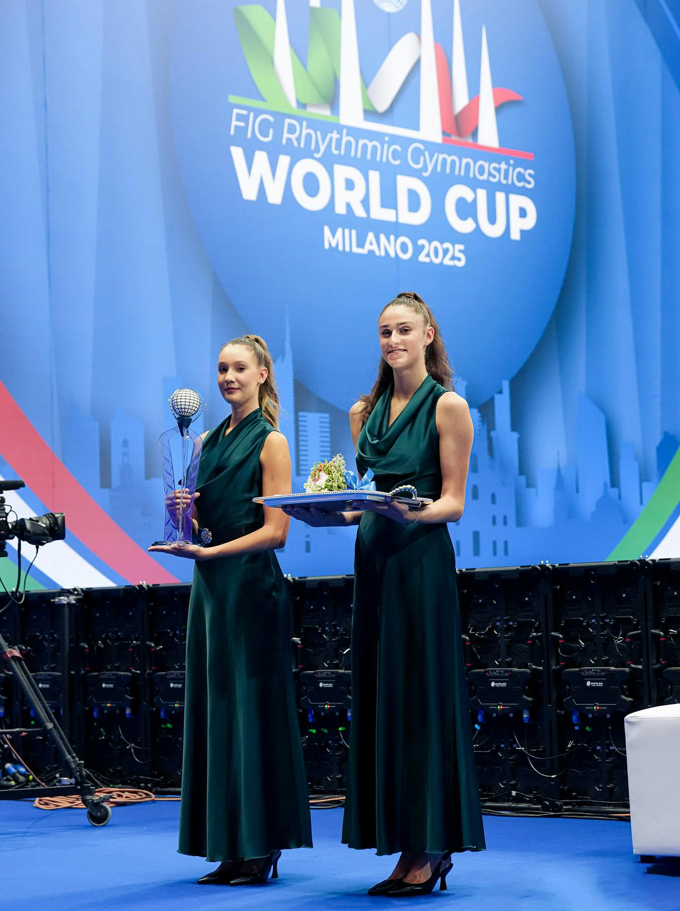 Two women in elegant green dresses stand on a blue stage at the FIG Rhythmic Gymnastics World Cup in Milan 2025, holding trophies.