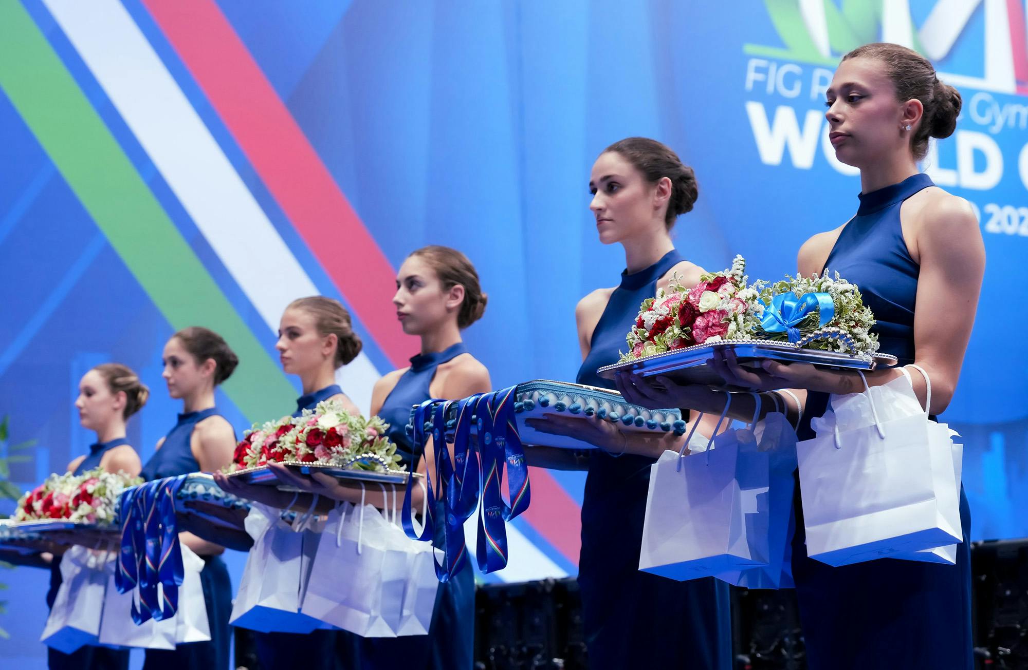 A group of women in blue dresses holds trays with medals and flower bouquets, standing against a blue backdrop with diagonal stripes. The scene conveys formality.