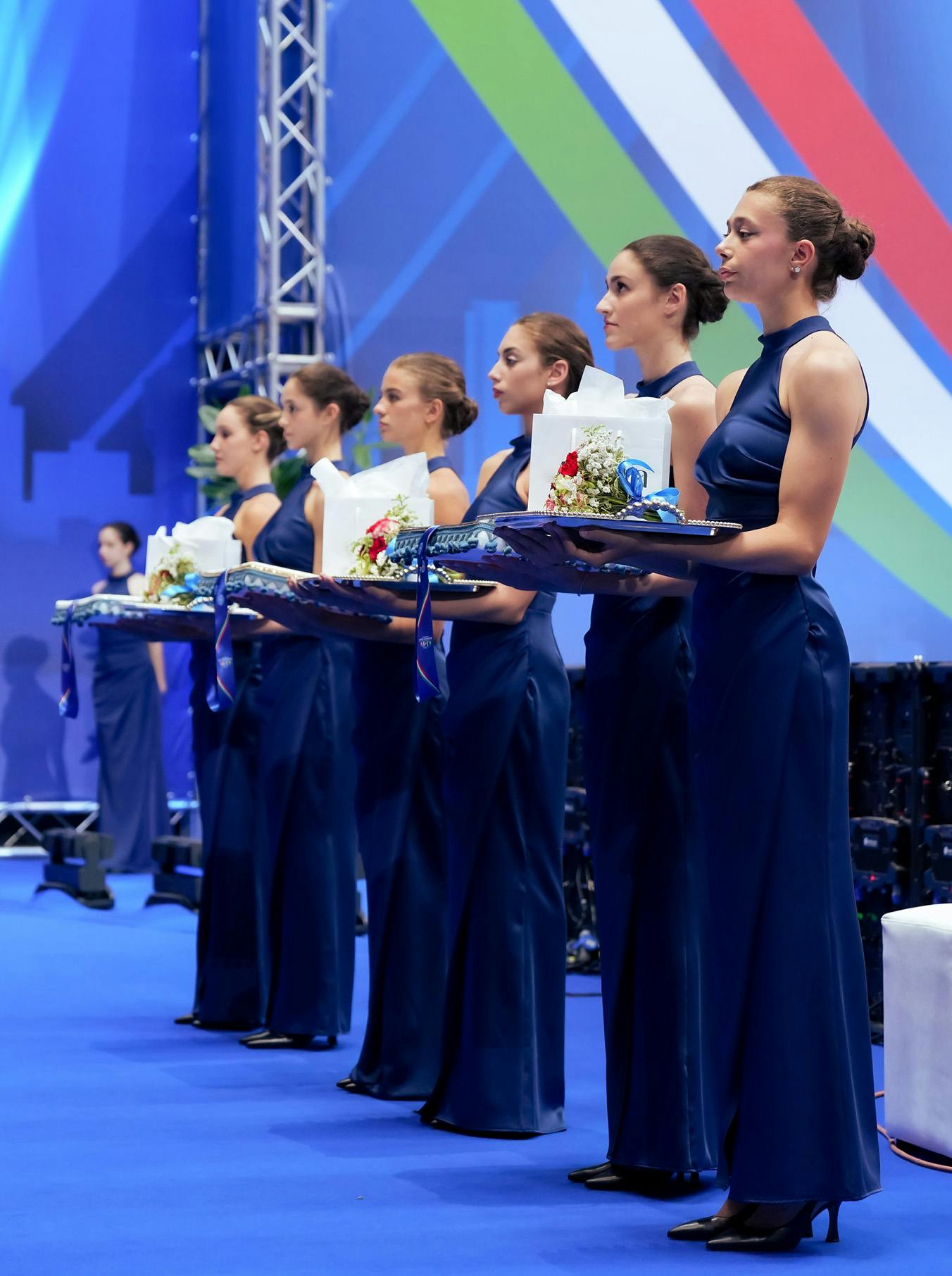Women in elegant blue gowns stand in a line, holding trays with wrapped gifts and flowers. The setting is formal, with a colorful backdrop.