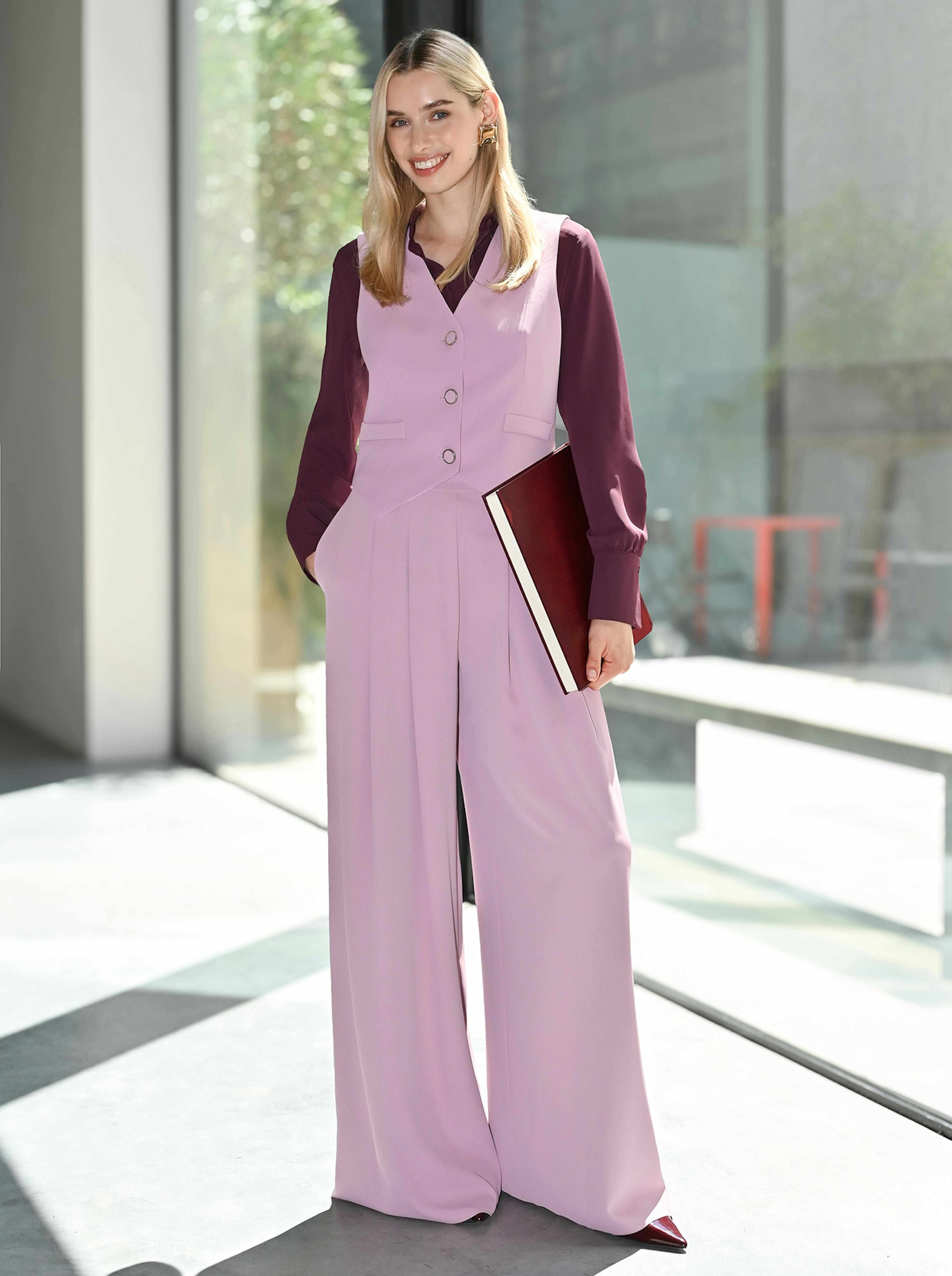 Smiling woman stands in sunlight wearing a mauve vest and wide-leg pants with a maroon blouse. She holds a book, exuding confidence and style.