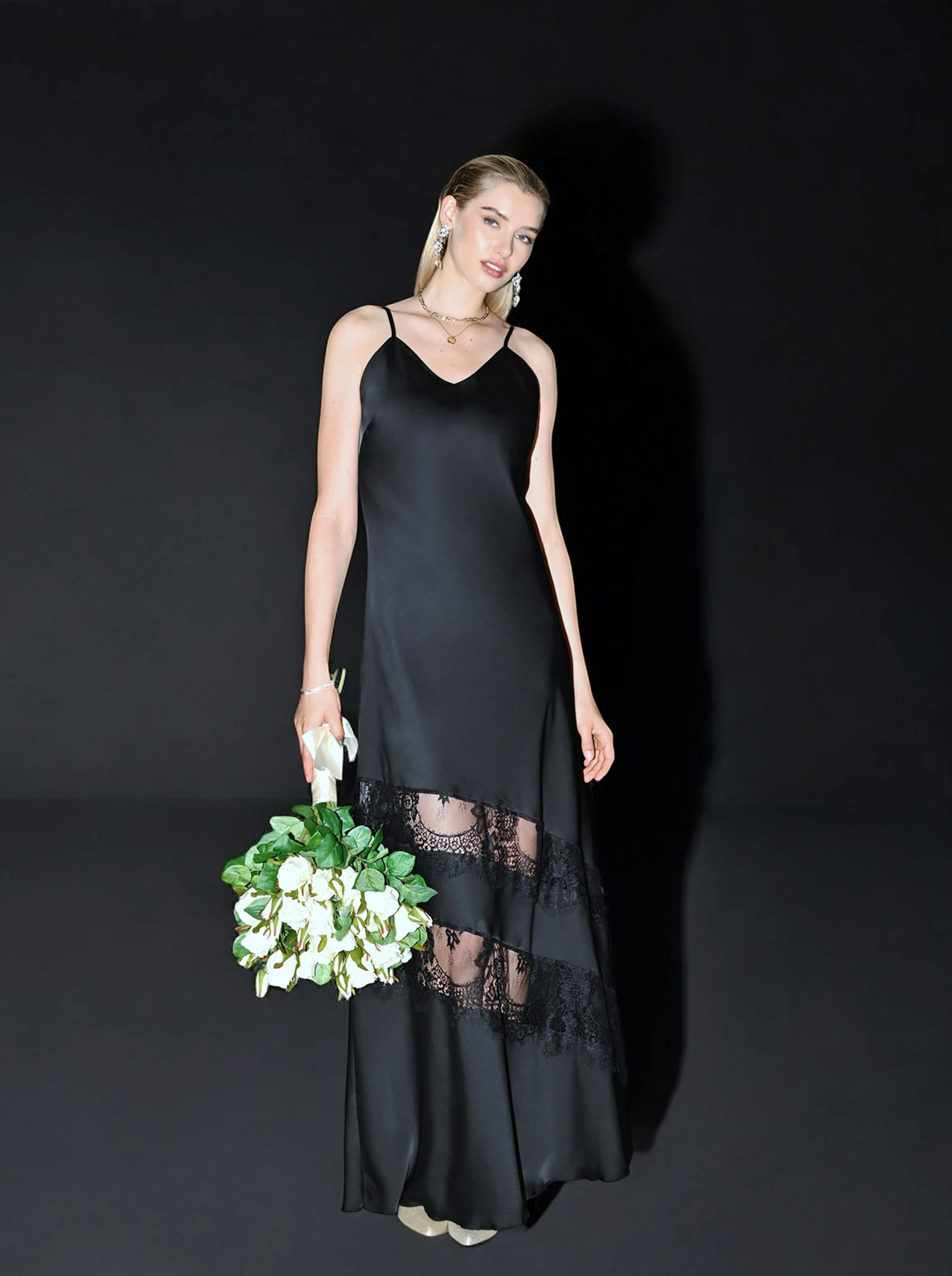 A woman in a sleek black dress with lace panels stands against a dark background, holding a bouquet of white and green flowers, exuding elegance.