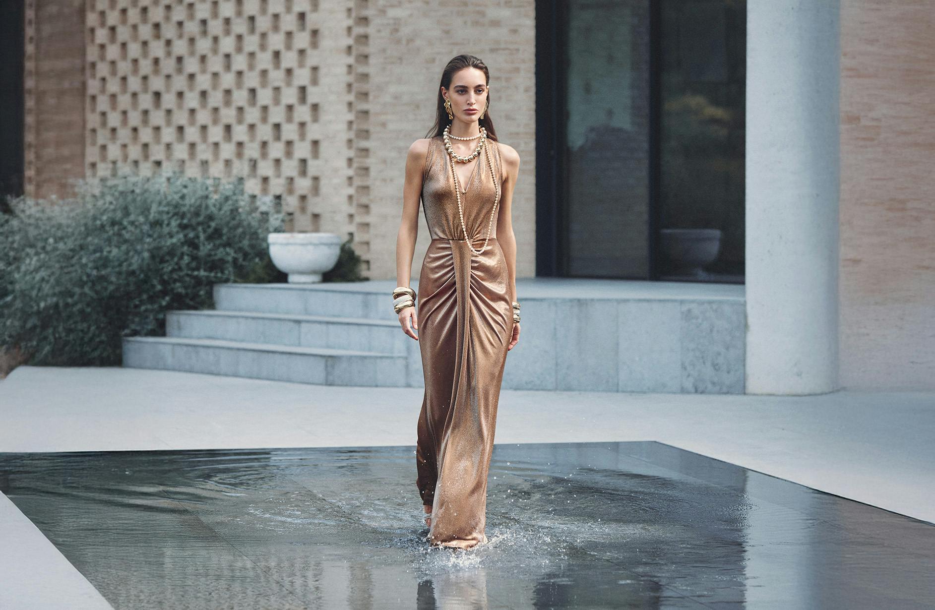Elegant woman in a shimmering gold gown walks confidently through water. Dramatic setting with modern architecture, exuding sophistication and style.