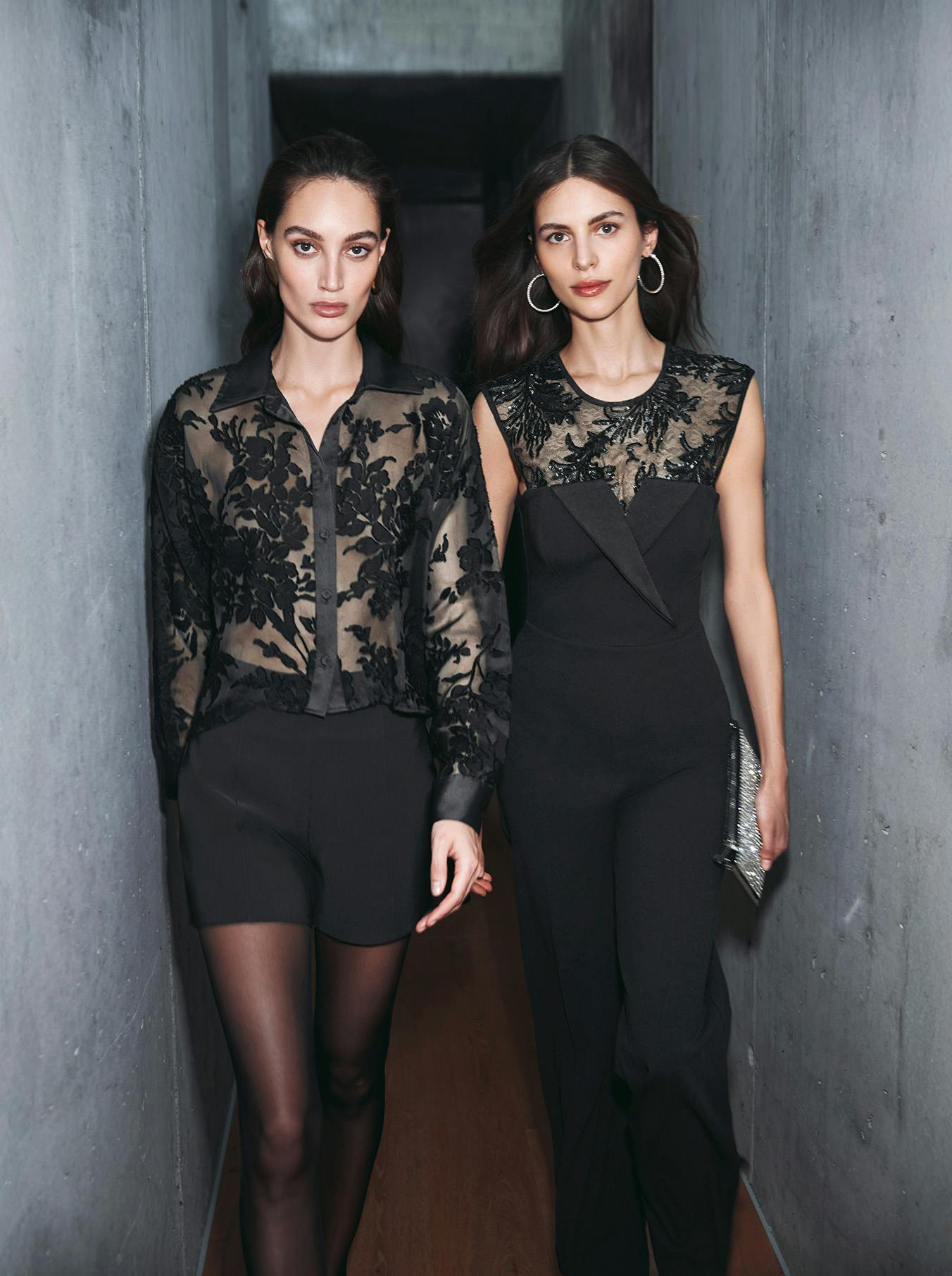 Two women confidently walk down a narrow hallway, both wearing elegant black outfits with lace details. The atmosphere is chic and sophisticated.