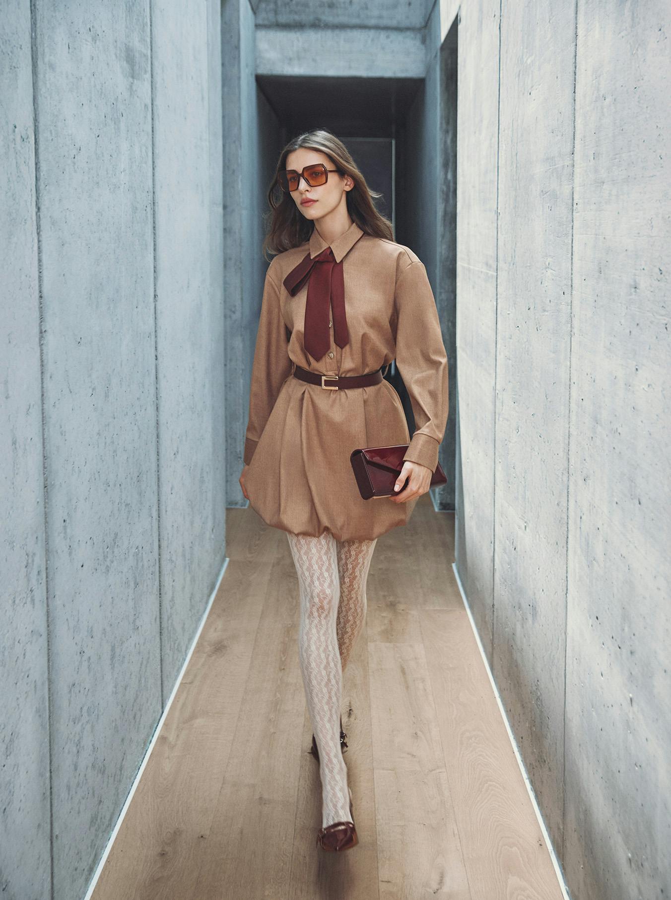 Woman in a tan dress with a burgundy scarf walks confidently through a concrete corridor. She wears oversized sunglasses, lace tights, and holds a clutch.