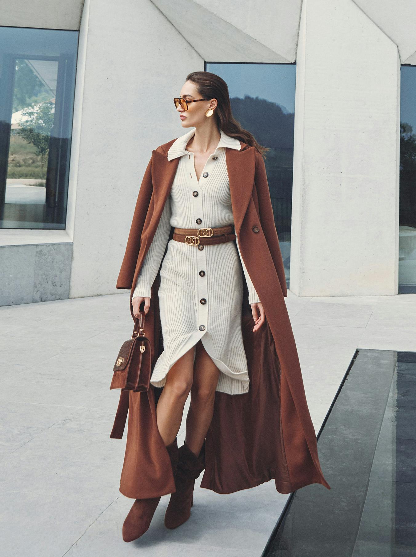 A stylish woman in sunglasses wearing a cream ribbed dress and brown coat walks confidently. She carries a brown handbag; the mood is elegant and poised.