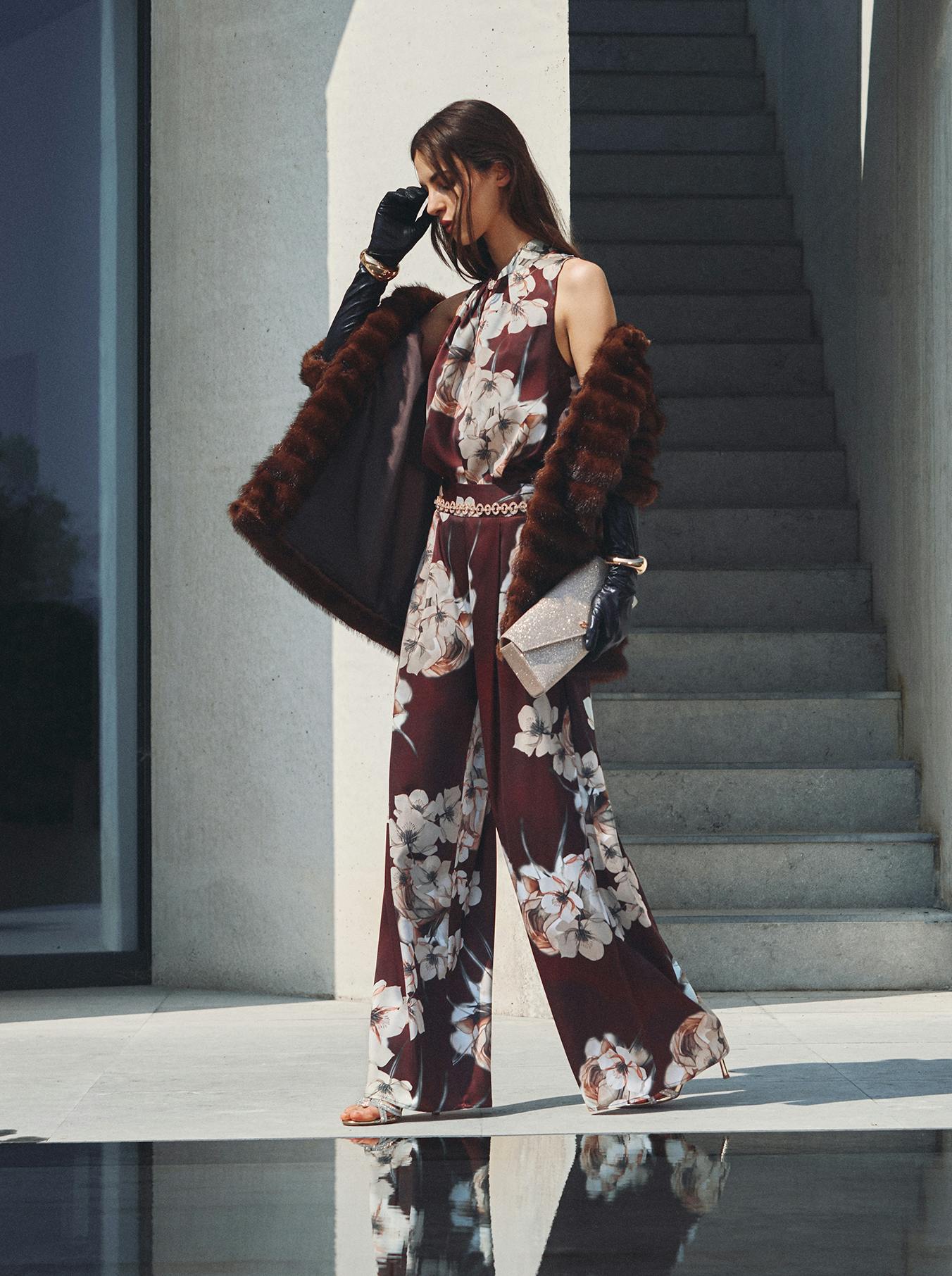 A woman in a floral jumpsuit and fur coat stands elegantly by a modern building. She holds a sparkly clutch and wears black gloves, conveying sophistication.