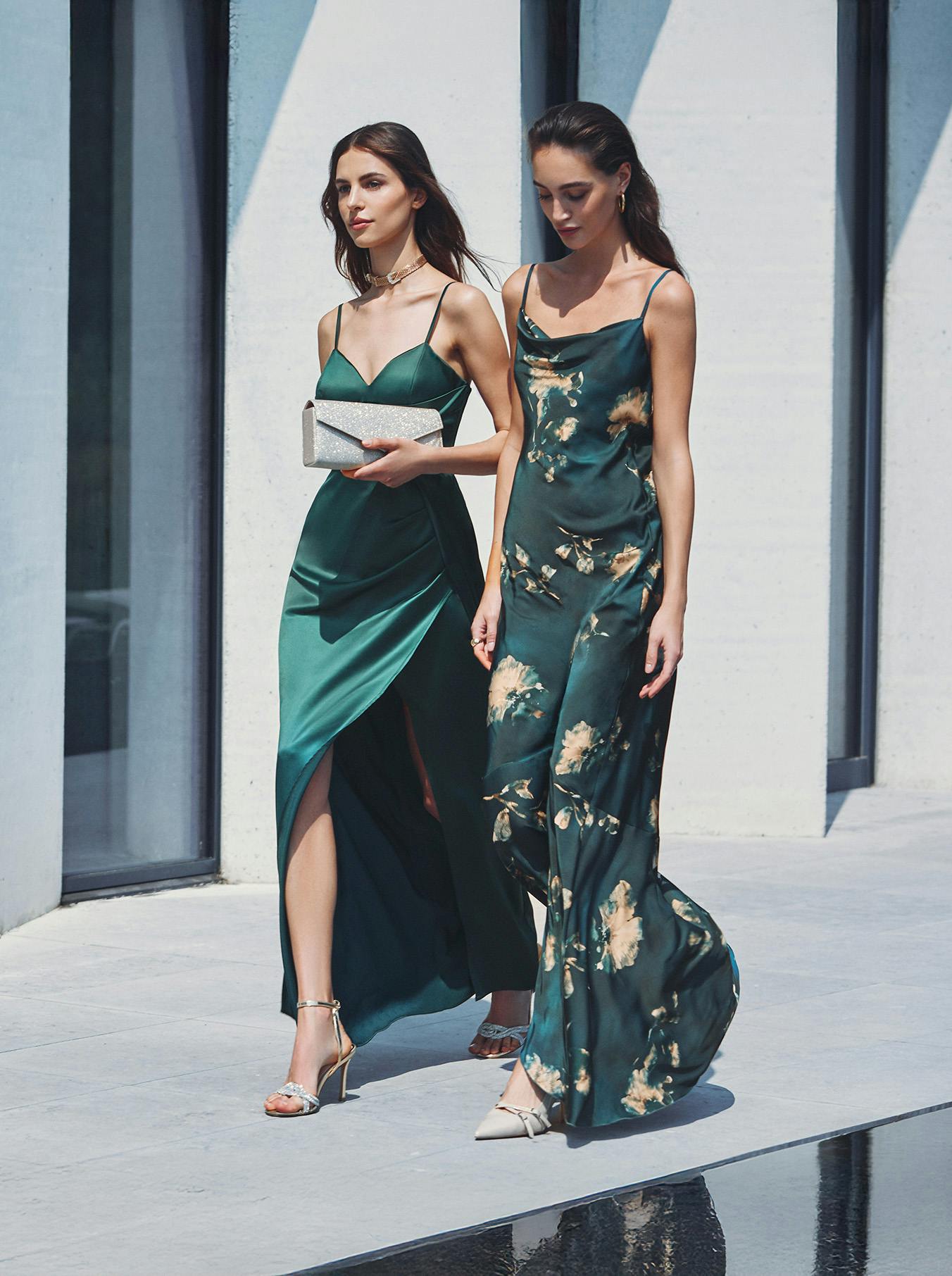 Two women in elegant green dresses walk confidently by a modern building. One holds a sparkling clutch. The scene conveys sophistication and style.
