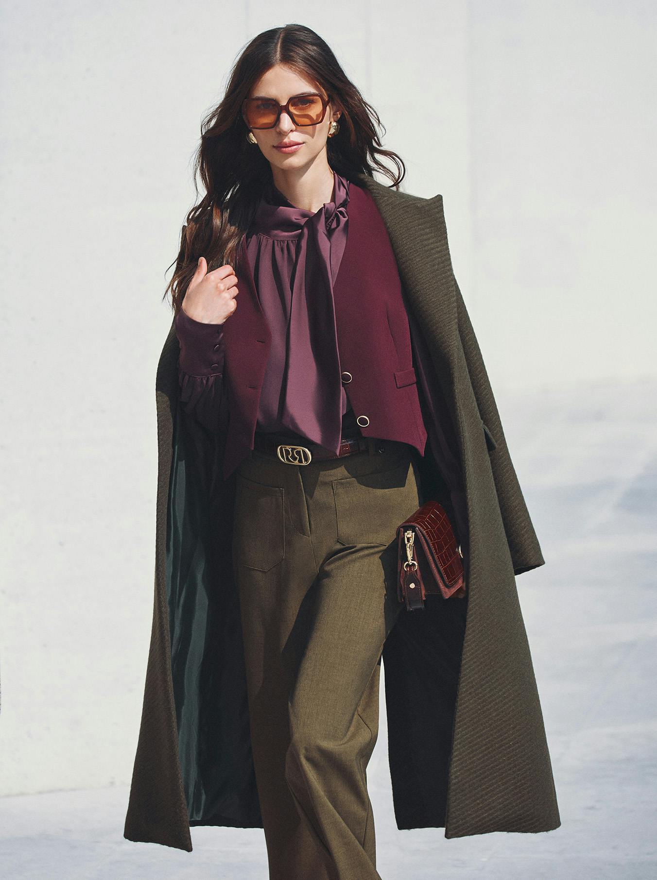 Fashionable woman walking confidently outdoors. She wears a dark green coat, burgundy blouse, olive trousers, and sunglasses, carrying a clutch. Elegant, stylish vibe.