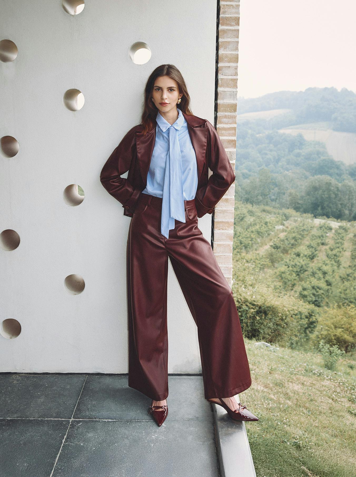 A woman in a maroon outfit and blue blouse leans against a wall with circular cutouts. Background shows rolling hills, creating a serene vibe.