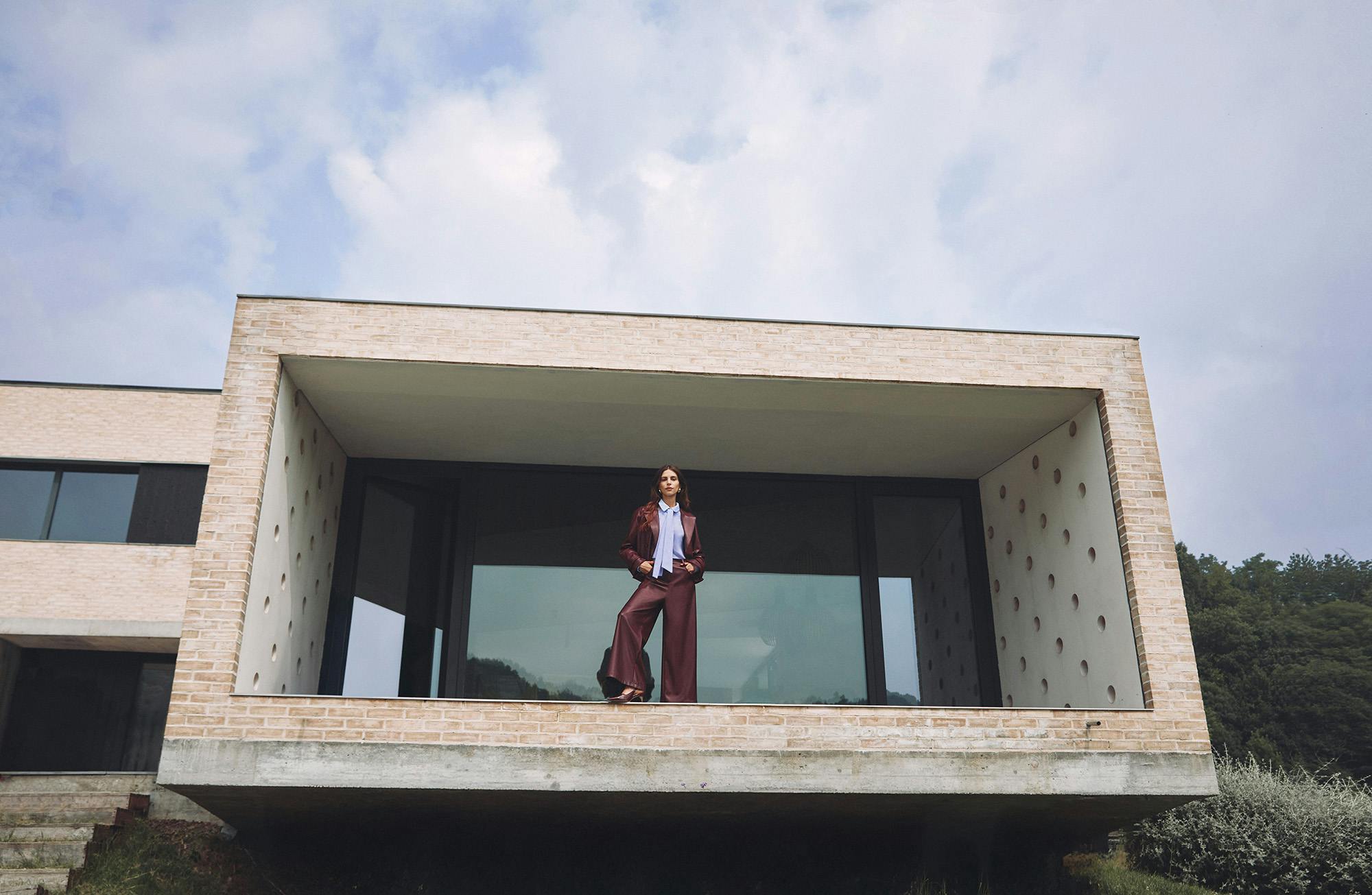 Woman in fashionable attire stands confidently on the balcony of a modern, rectangular building with brick and glass elements, under a cloudy sky.