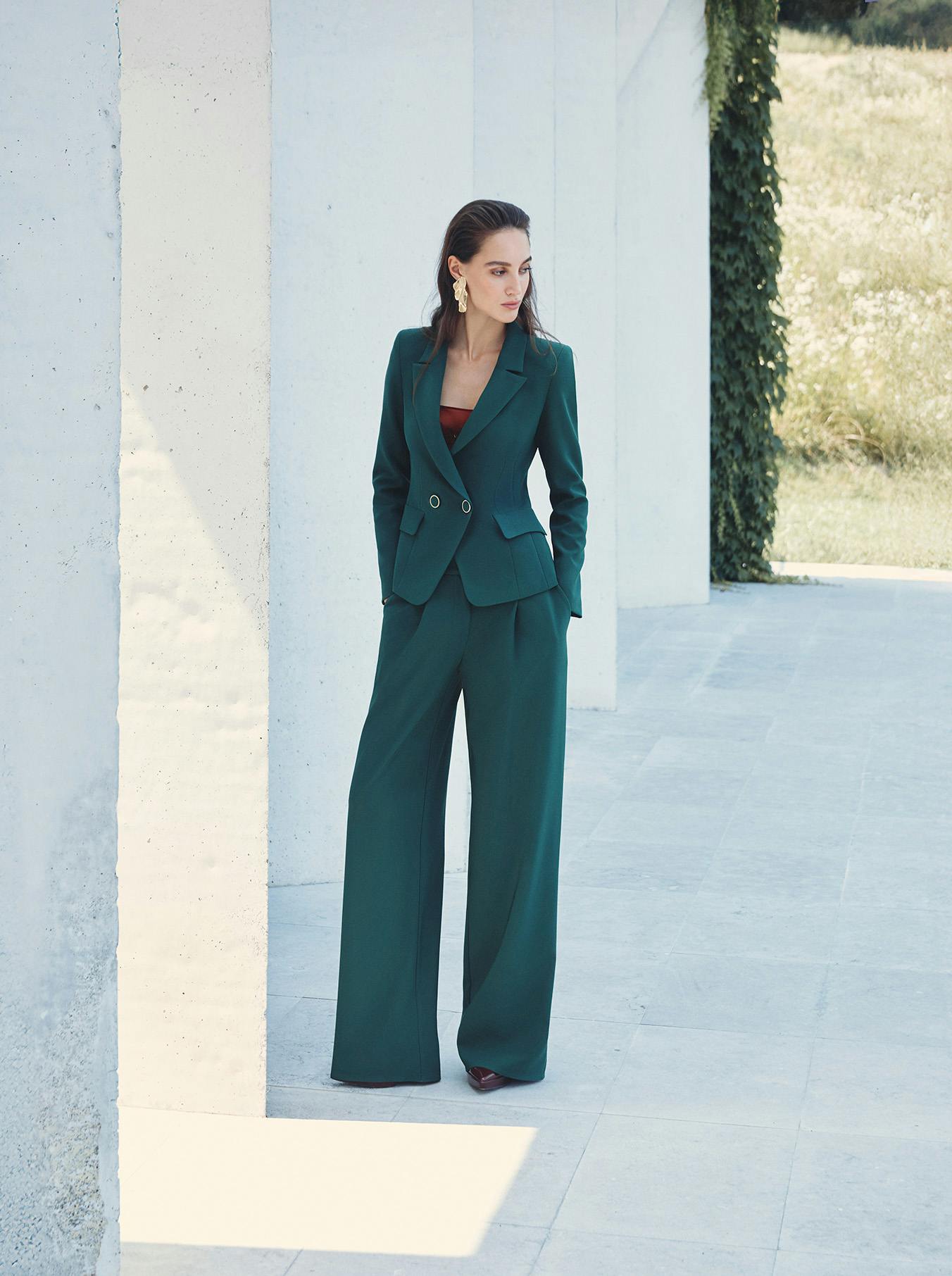 A woman in a dark teal pantsuit stands outdoors next to white pillars. Her expression is serene. Background reveals greenery and bright sunlight.