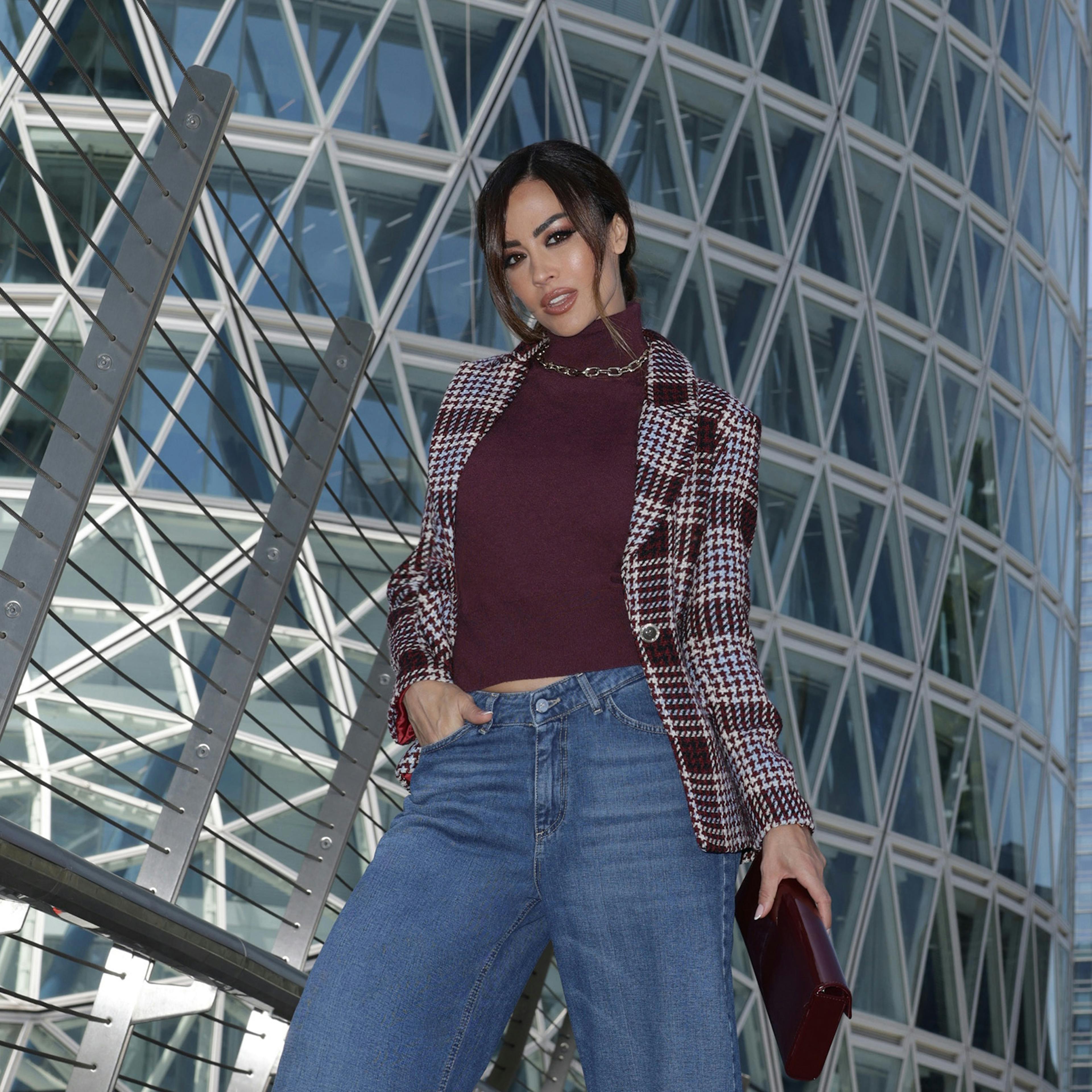Giorgia Palmas in wide-leg jeans and plaid jacket stands confidently on urban walkway. Futuristic skyscrapers with glass facades rise in the background.