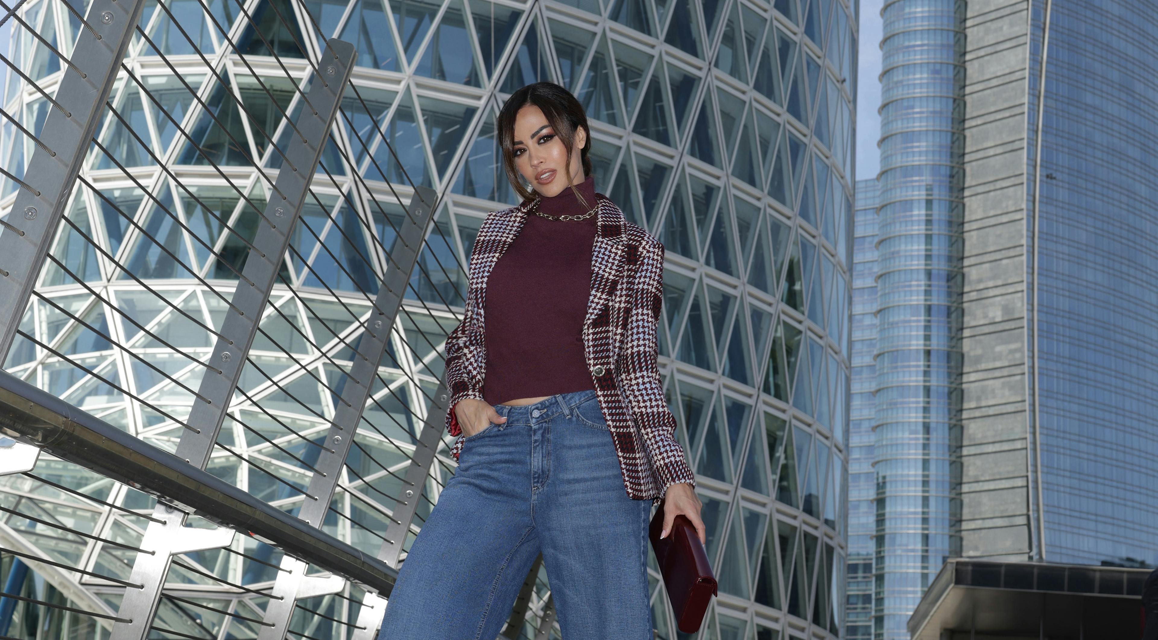 Giorgia Palmas in wide-leg jeans and plaid jacket stands confidently on urban walkway. Futuristic skyscrapers with glass facades rise in the background.