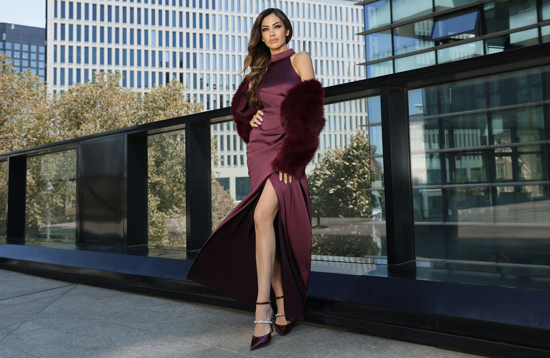 Giorgia Palmas in a maroon gown with a thigh-high slit and fur wrap stands confidently outdoors. She is on a city balcony, with modern glass buildings behind her.