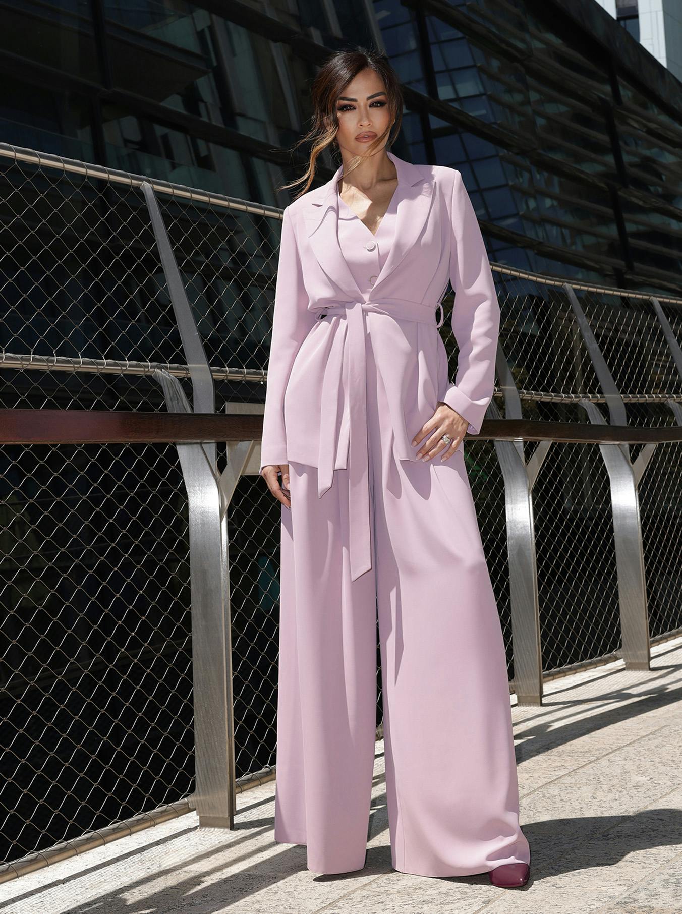 Giorgia Palmas in a stylish, light purple suit poses confidently outdoors. She stands on a sunlit stone path with a modern glass building in the background.
