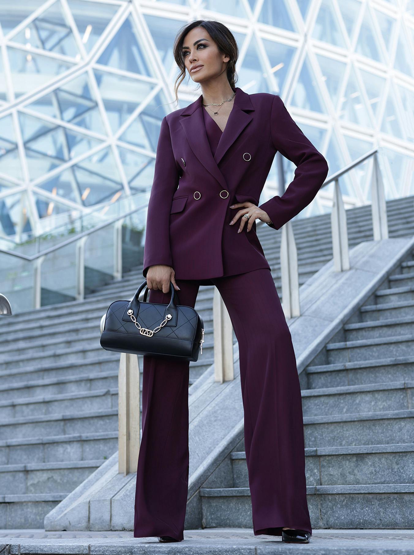 Giorgia Palmas in a stylish purple suit poses confidently on modern architectural stairs, holding a black purse. The background features a geometric glass structure.