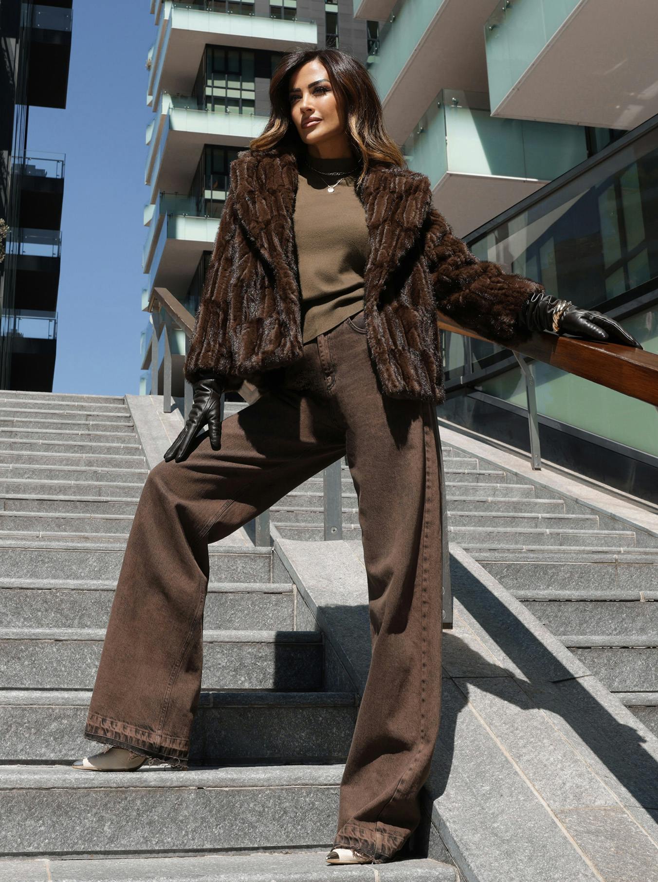 Giorgia Palmas in brown faux fur coat and wide-legged pants stands confidently on outdoor stairs. Modern building behind, conveying urban chic style.