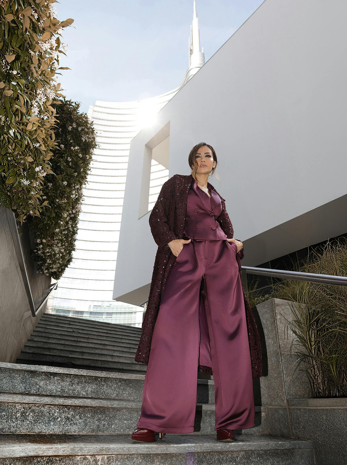 Giorgia Palmas in a burgundy suit and coat stands confidently on stone steps, with a modern building and greenery framing the scene. Bright and stylish ambiance.