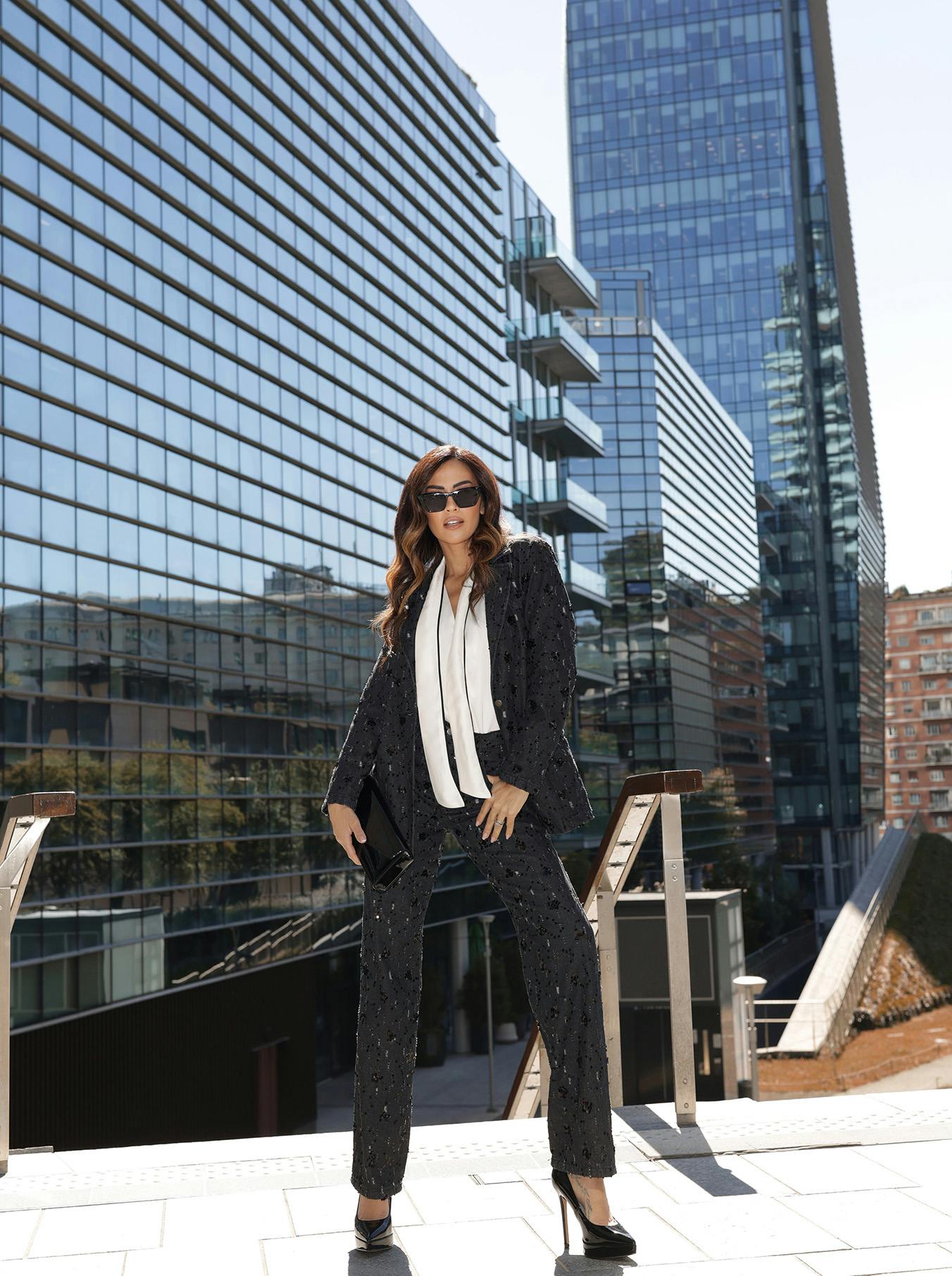 Giorgia Plamas in a stylish black suit and white blouse stands confidently in front of modern glass buildings, wearing sunglasses and holding a clutch. Elegant urban setting.