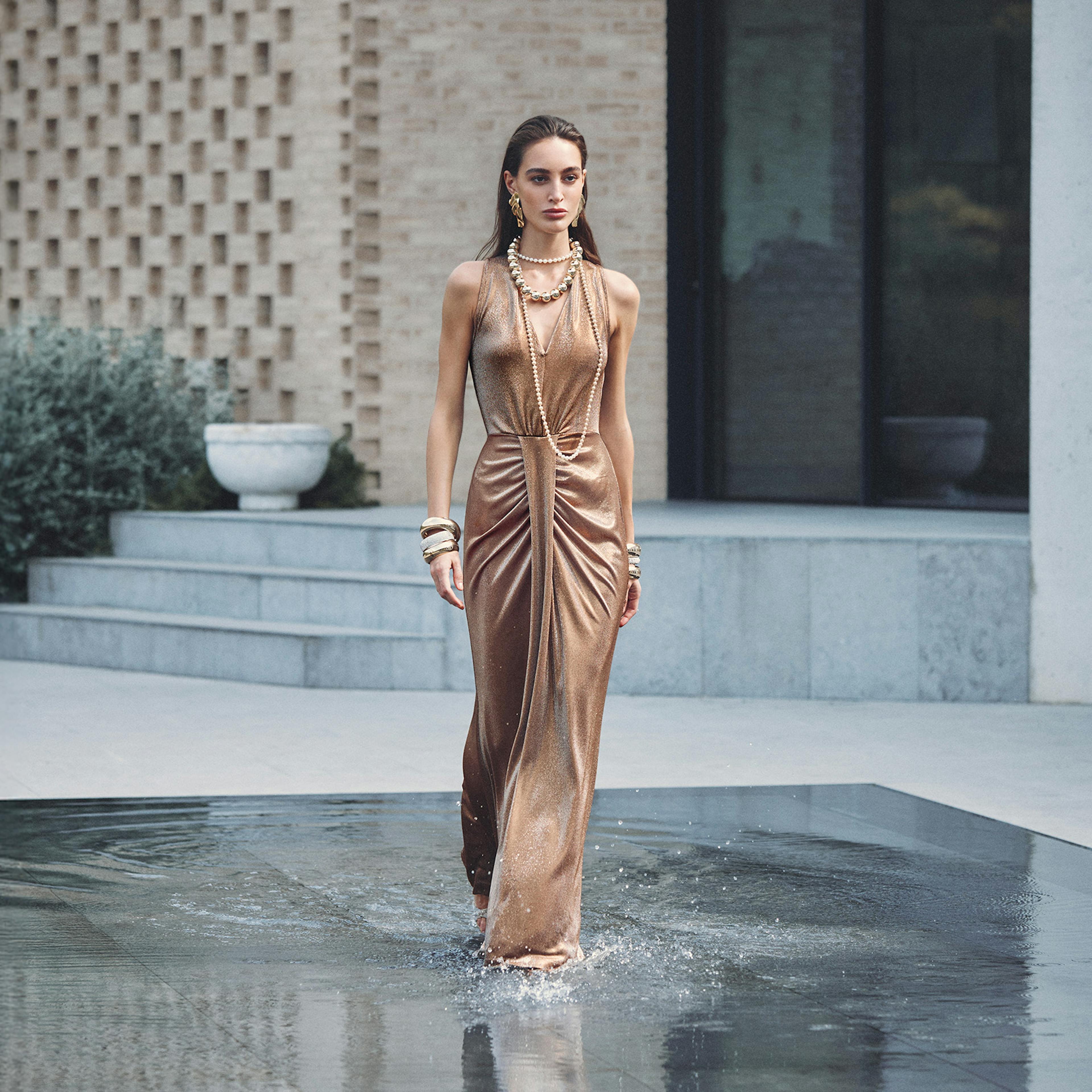 A model in a shimmering metallic gown walks on a reflective wet runway surrounded by modern architecture and greenery, exuding elegance and sophistication.