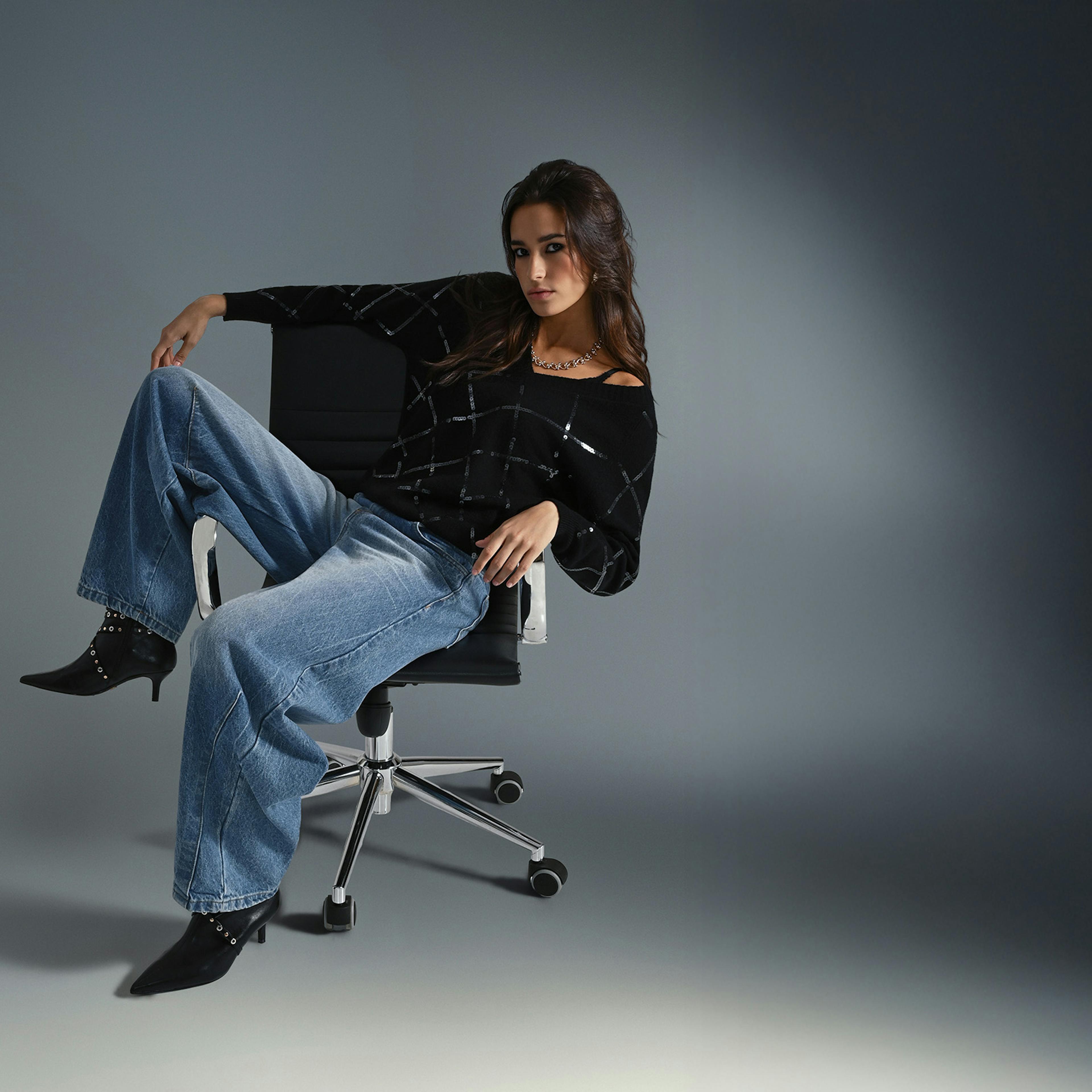 A woman sits relaxed in an office chair against a dark background. She wears a black top and loose blue jeans, exuding a confident, casual vibe.