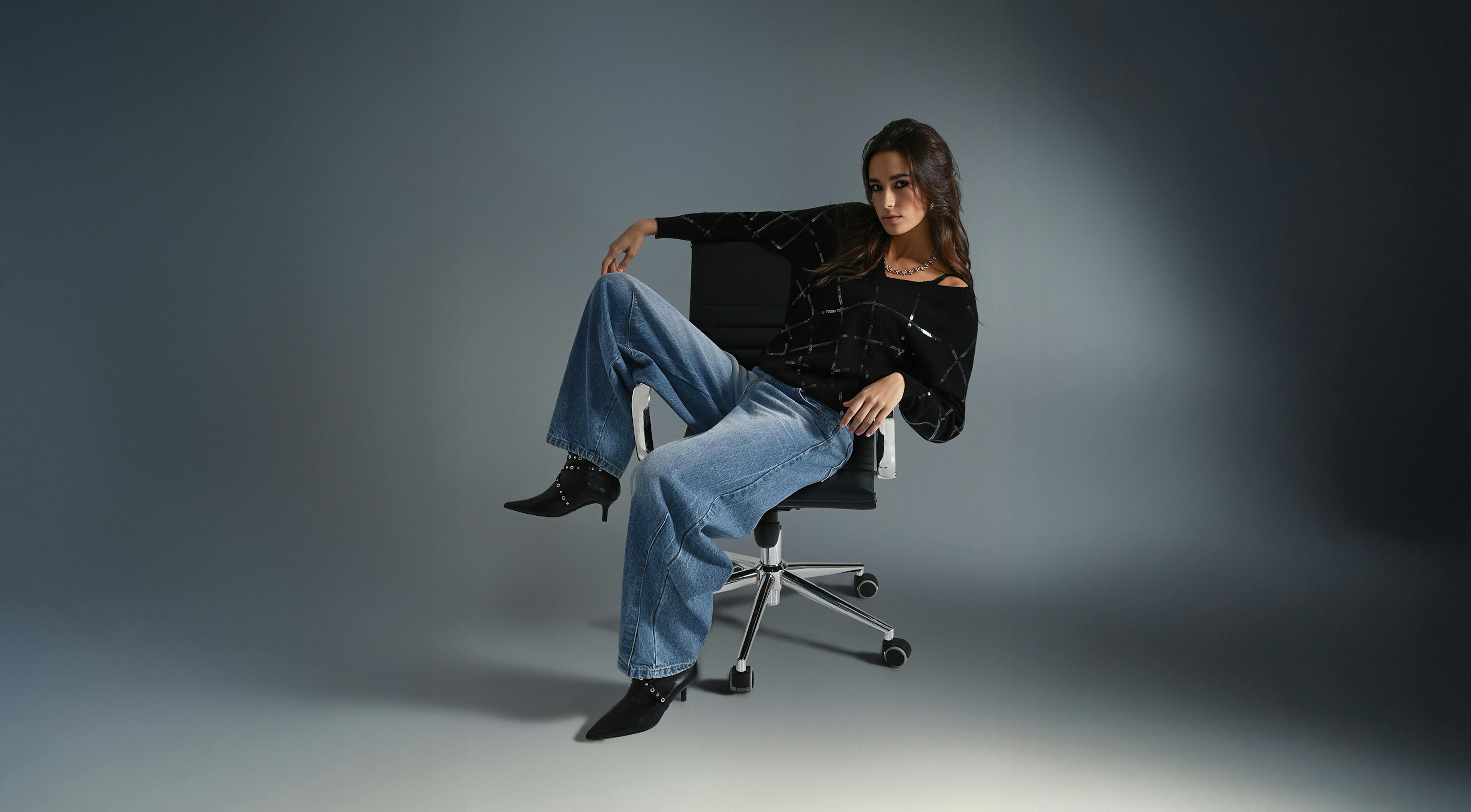 A woman sits relaxed in an office chair against a dark background. She wears a black top and loose blue jeans, exuding a confident, casual vibe.