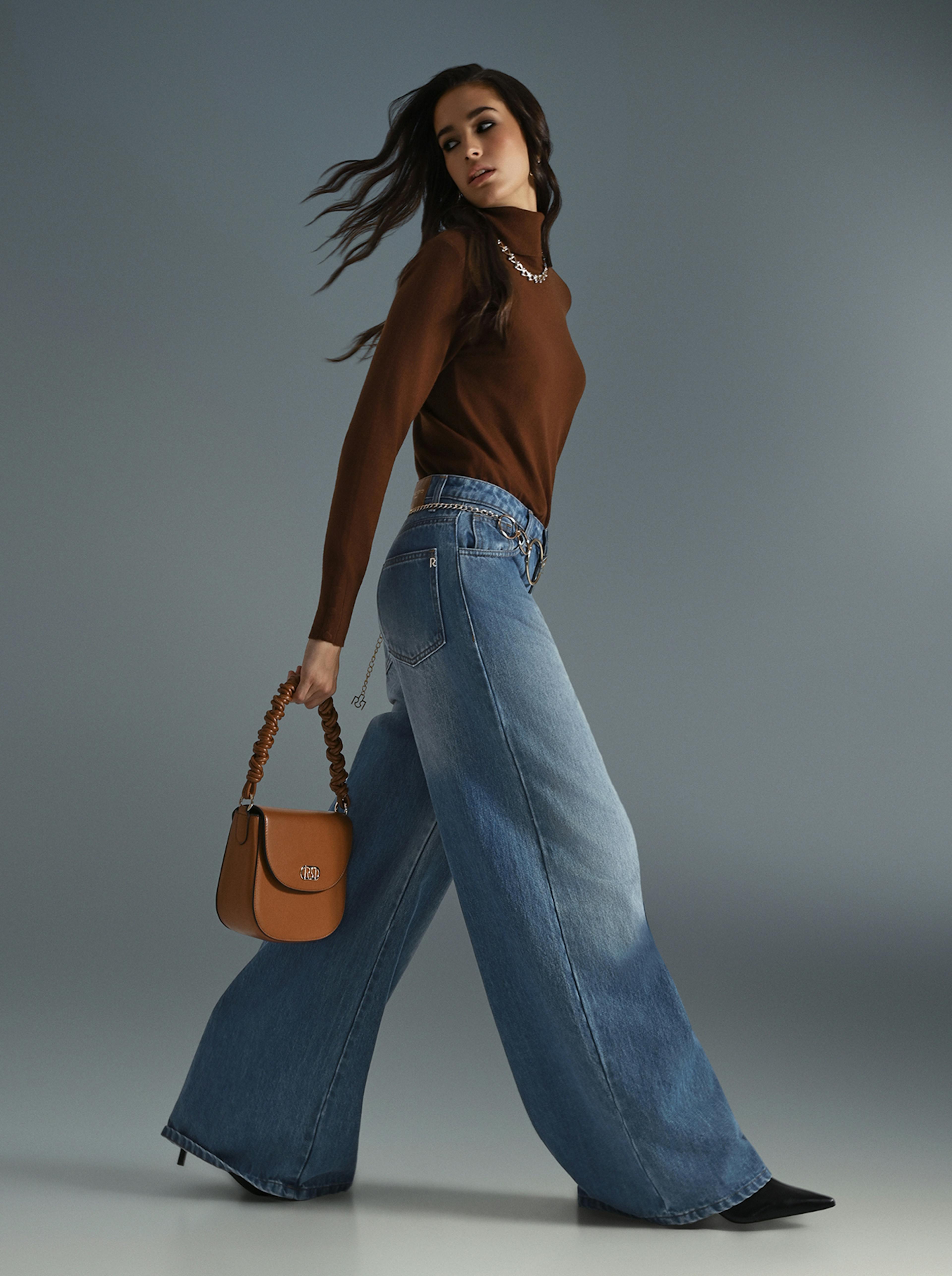 A woman in a brown sweater and jeans holds a bag, standing casually in an outdoor setting.