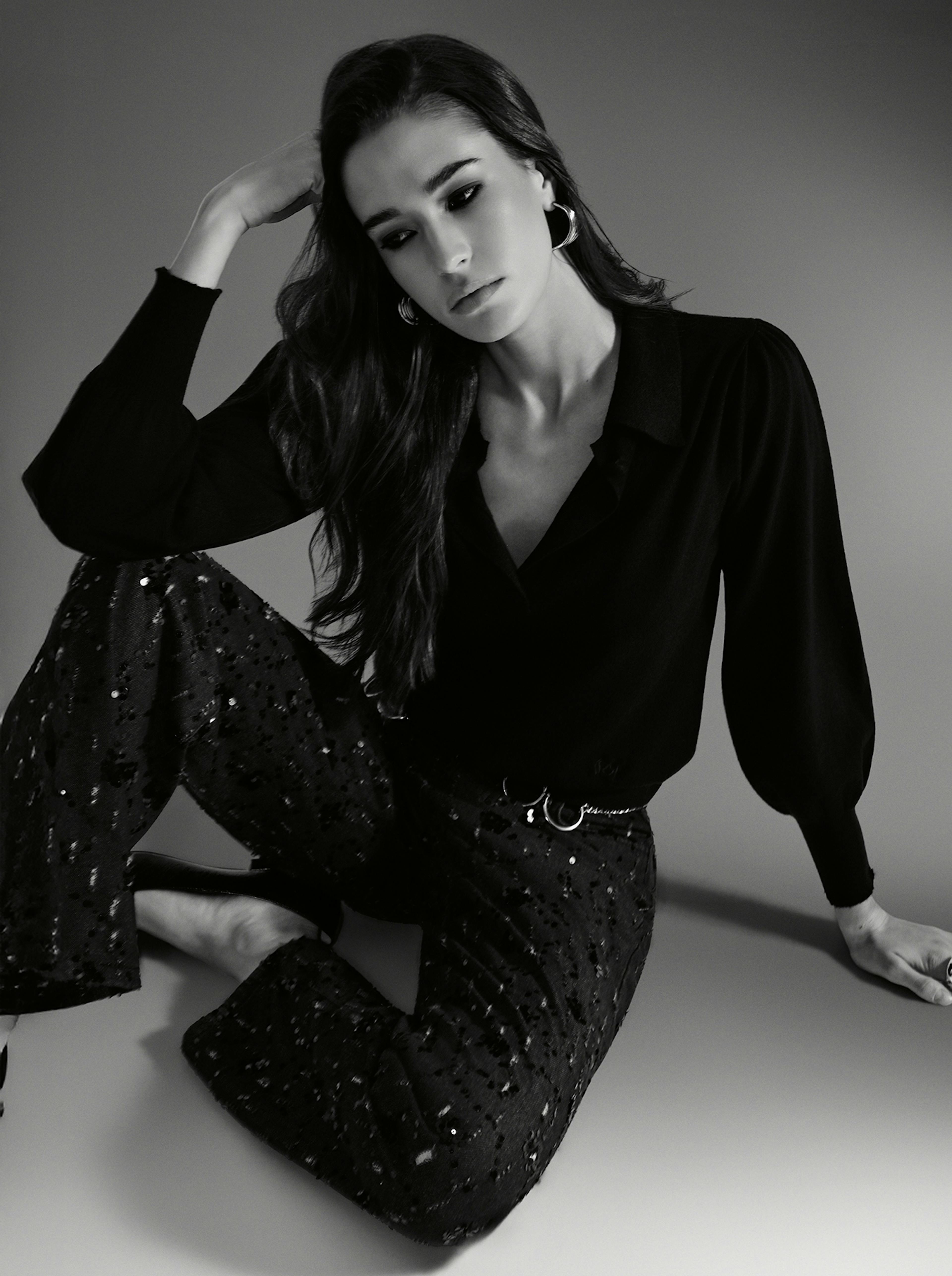 A woman in black and white denim sitting on the floor, looking contemplative and relaxed.