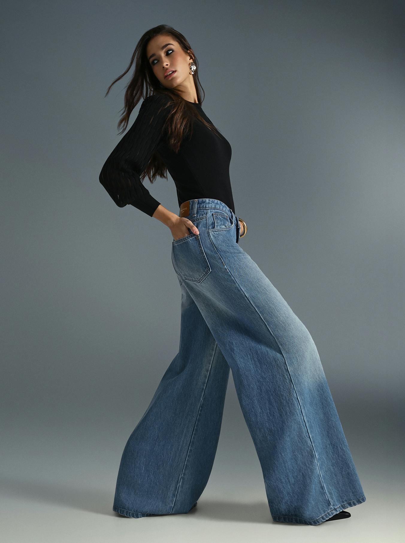 A woman wearing a black top and wide jeans stands confidently, showcasing a casual yet stylish outfit.