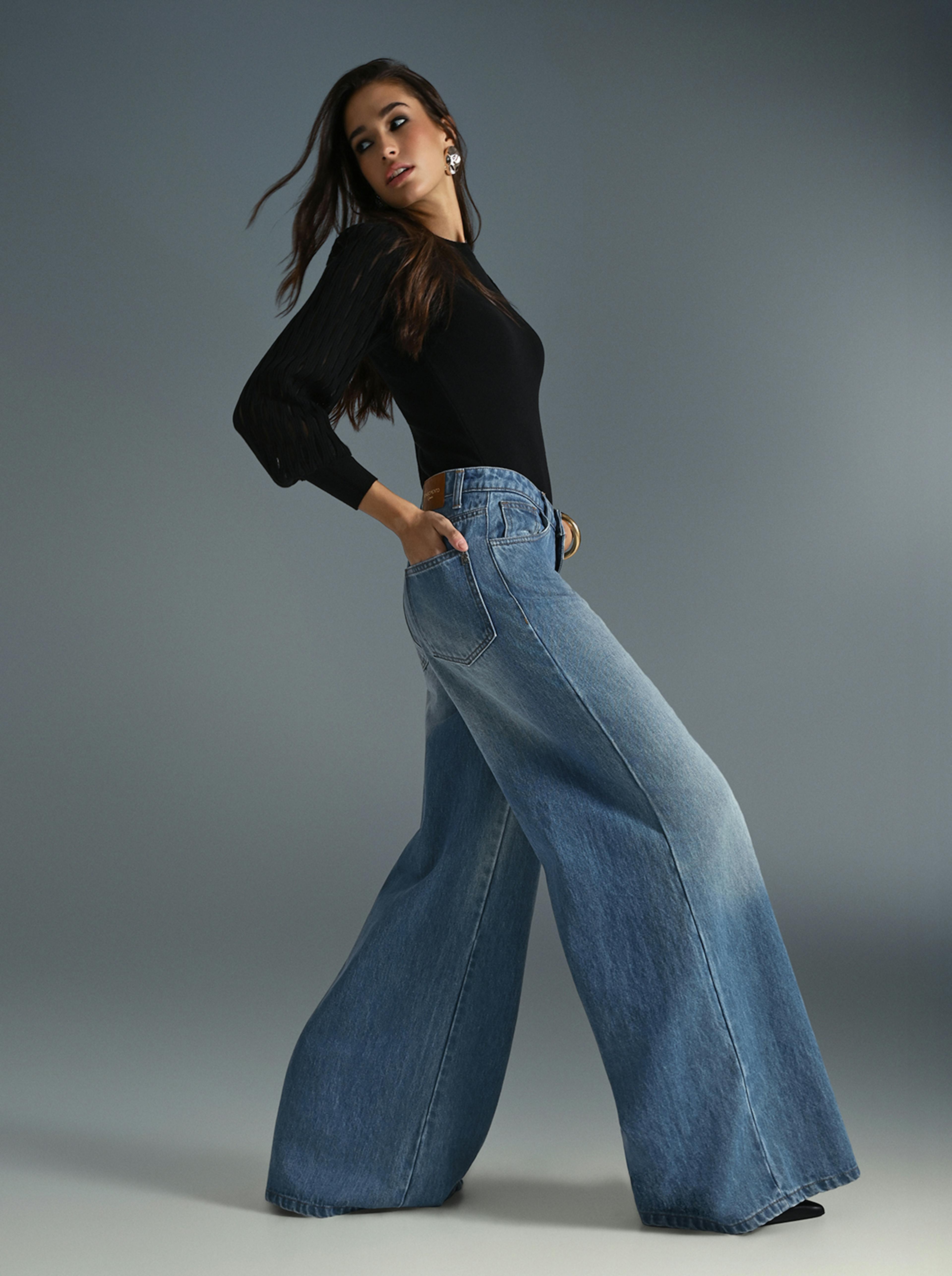 A woman wearing a black top and wide jeans stands confidently, showcasing a casual yet stylish outfit.