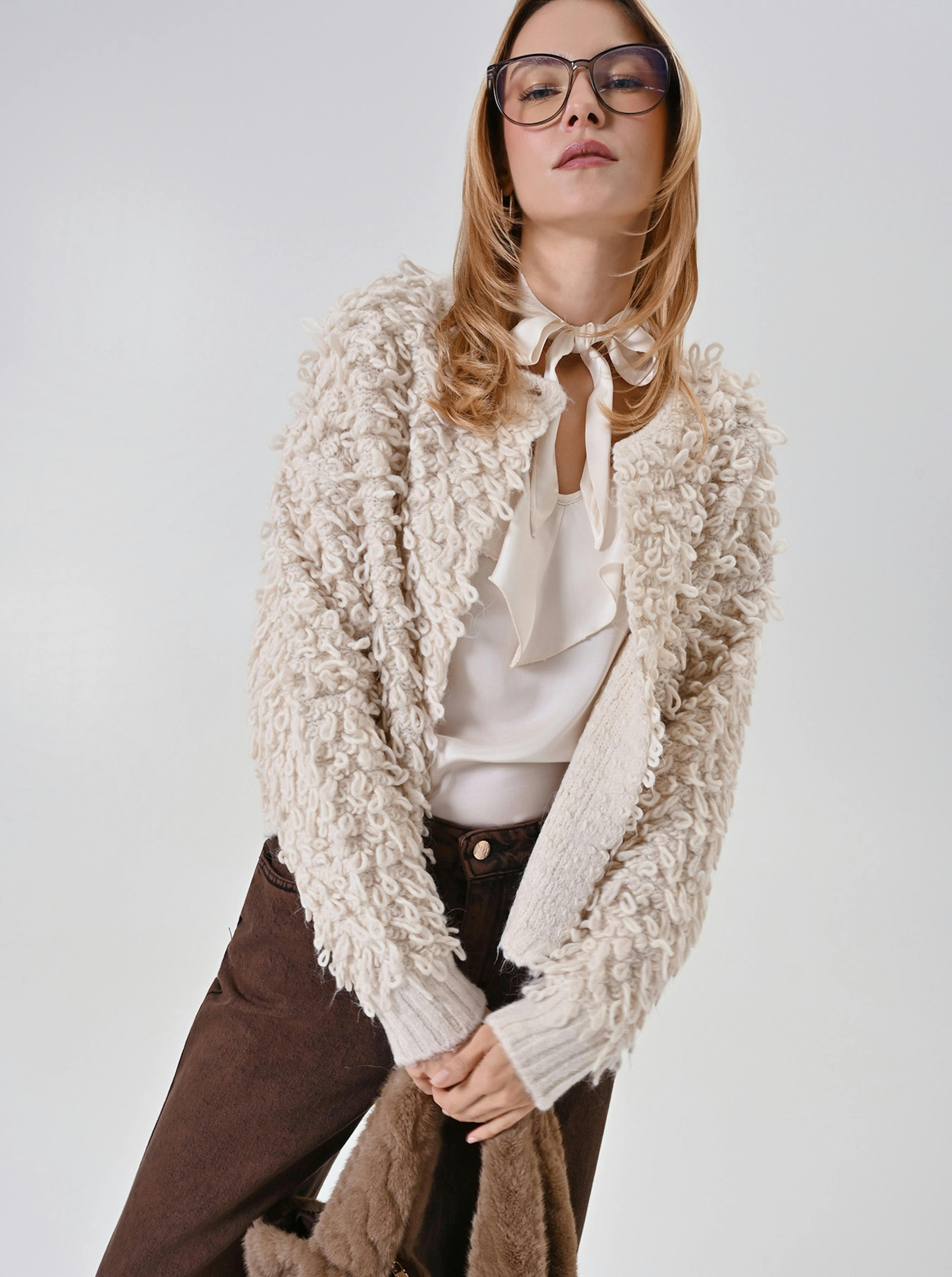 A person wearing glasses and a look of confidence poses in a textured cream sweater, white blouse, and brown pants, holding a fur coat in a relaxed, stylish manner.