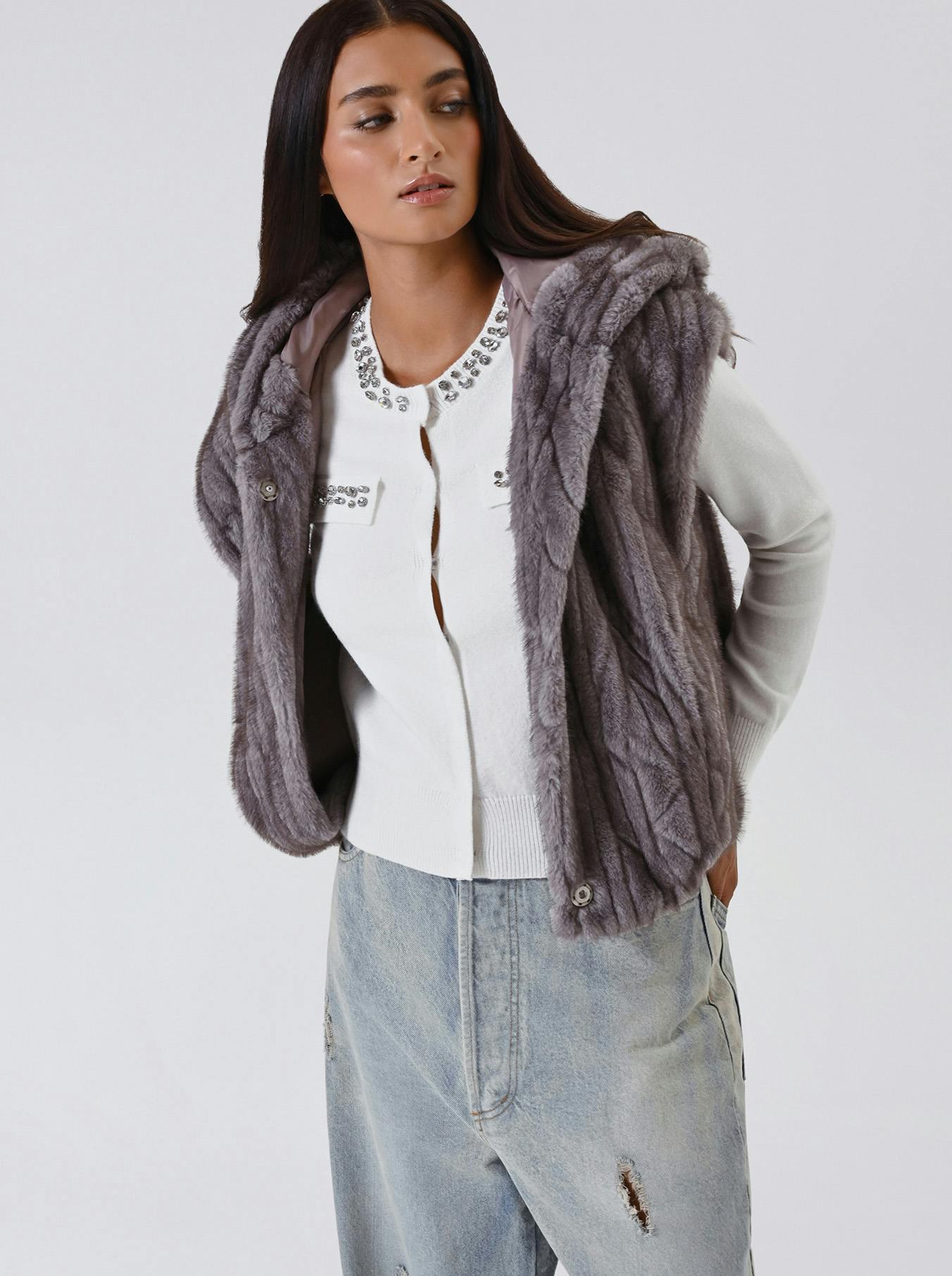 Woman with long dark hair wearing a gray faux fur vest over a white, embellished sweater and light-wash jeans. She poses confidently against a plain background.