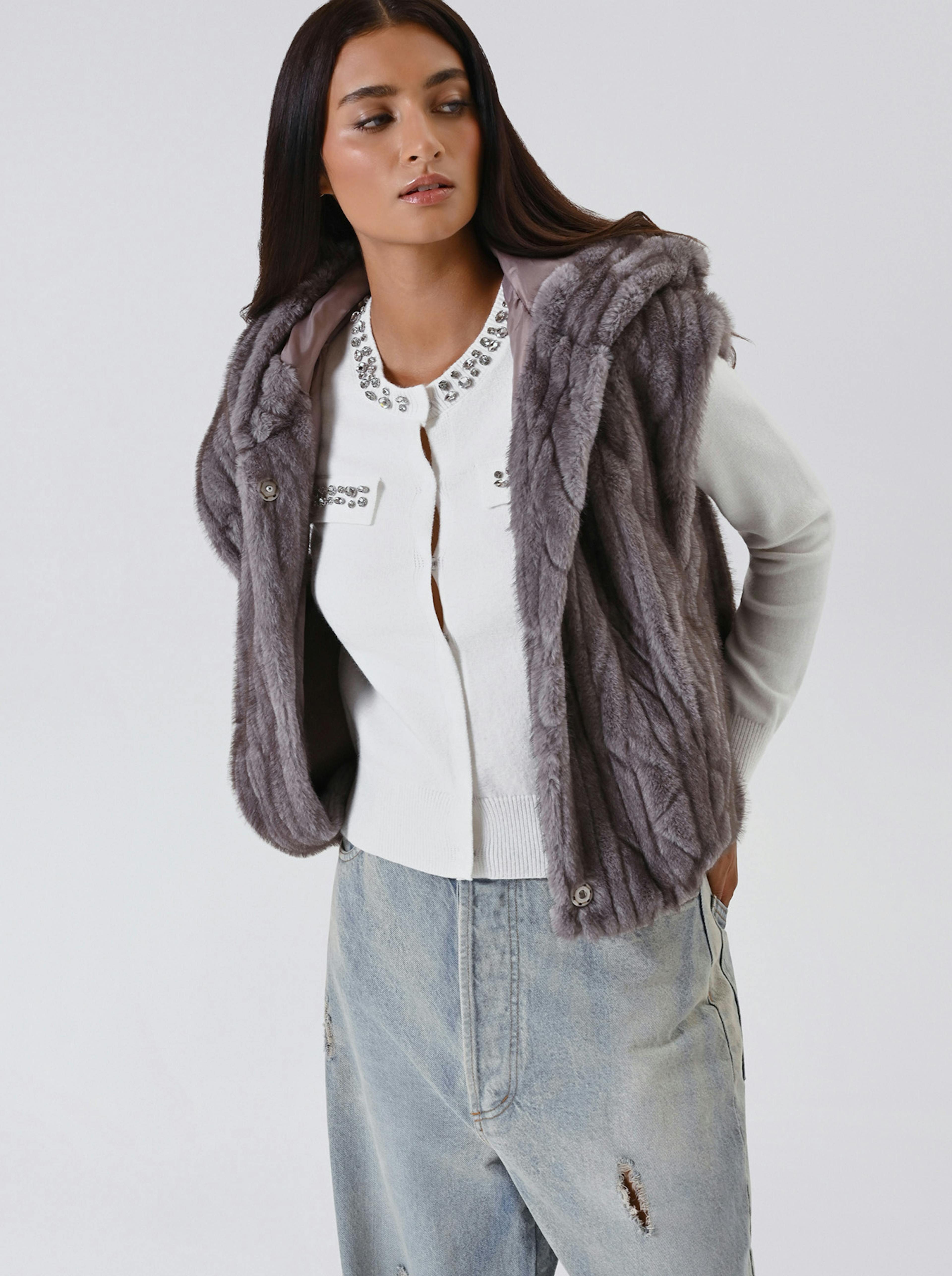 Woman with long dark hair wearing a gray faux fur vest over a white, embellished sweater and light-wash jeans. She poses confidently against a plain background.