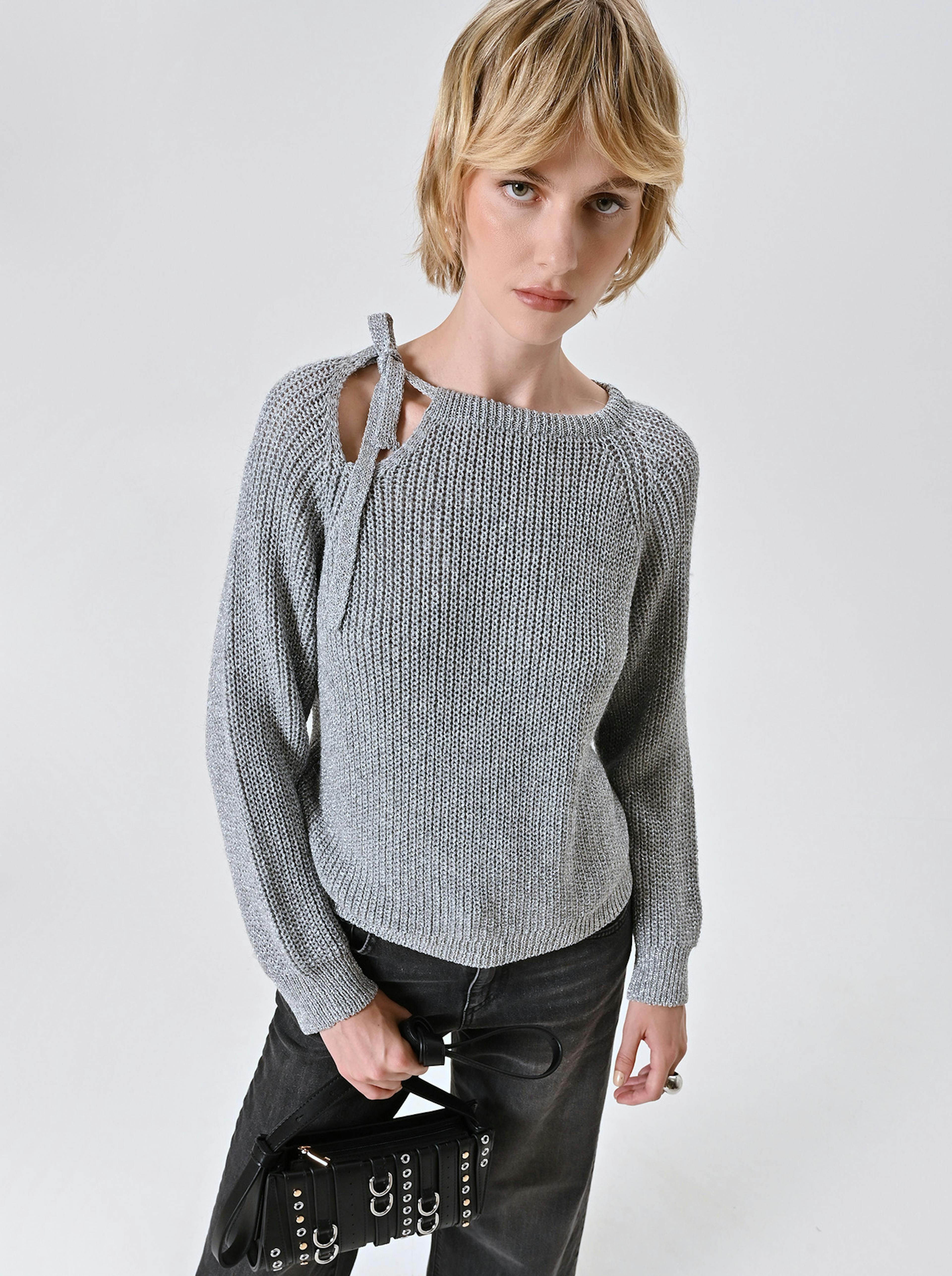 A person with short blonde hair wears a gray knit sweater with a cutout and bow on the shoulder, holding a black studded handbag. Their expression is serious.