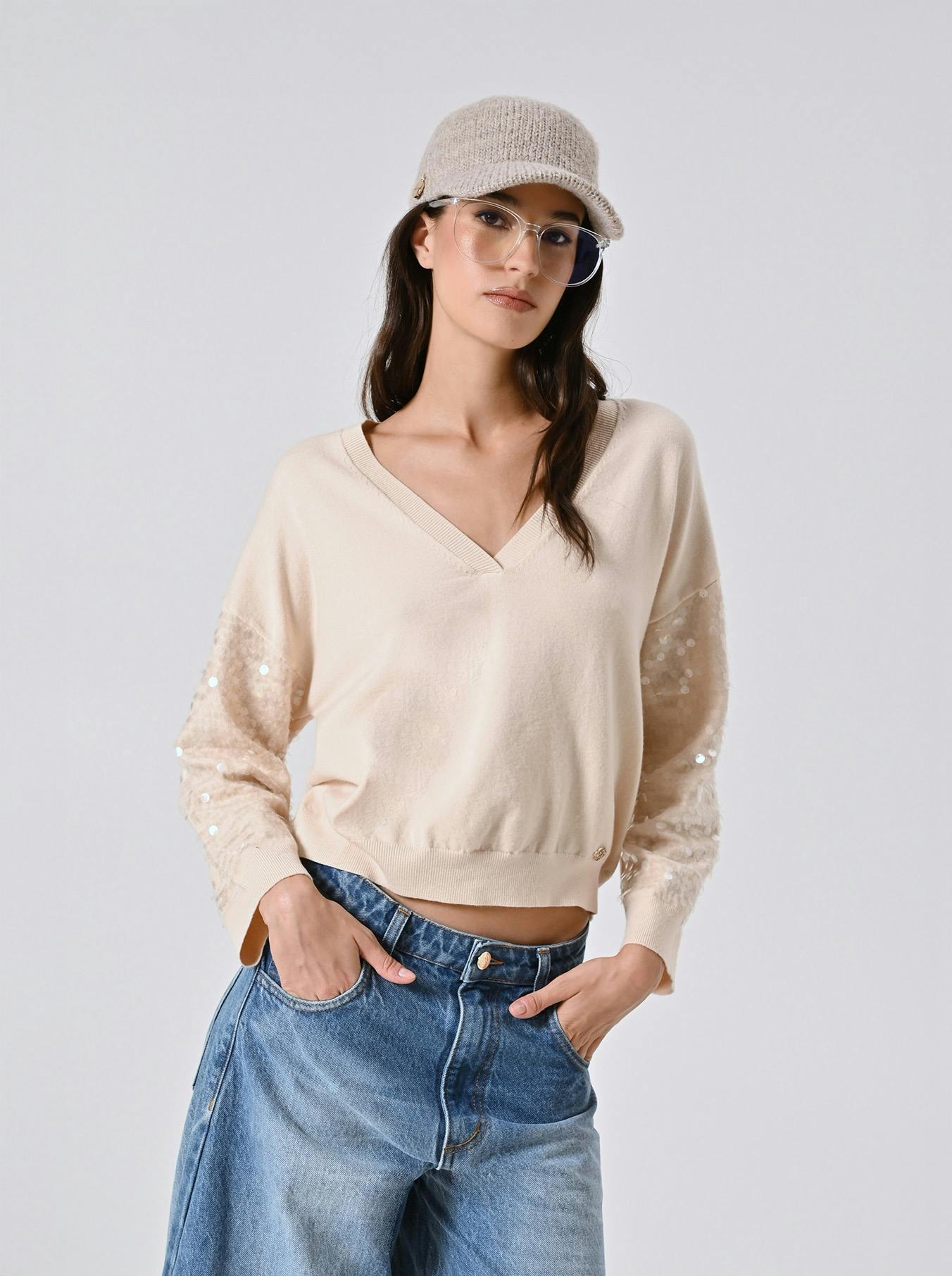 A woman in a beige sweater with sequin sleeves, blue jeans, and a knit cap poses confidently with hands in her pockets against a plain background.