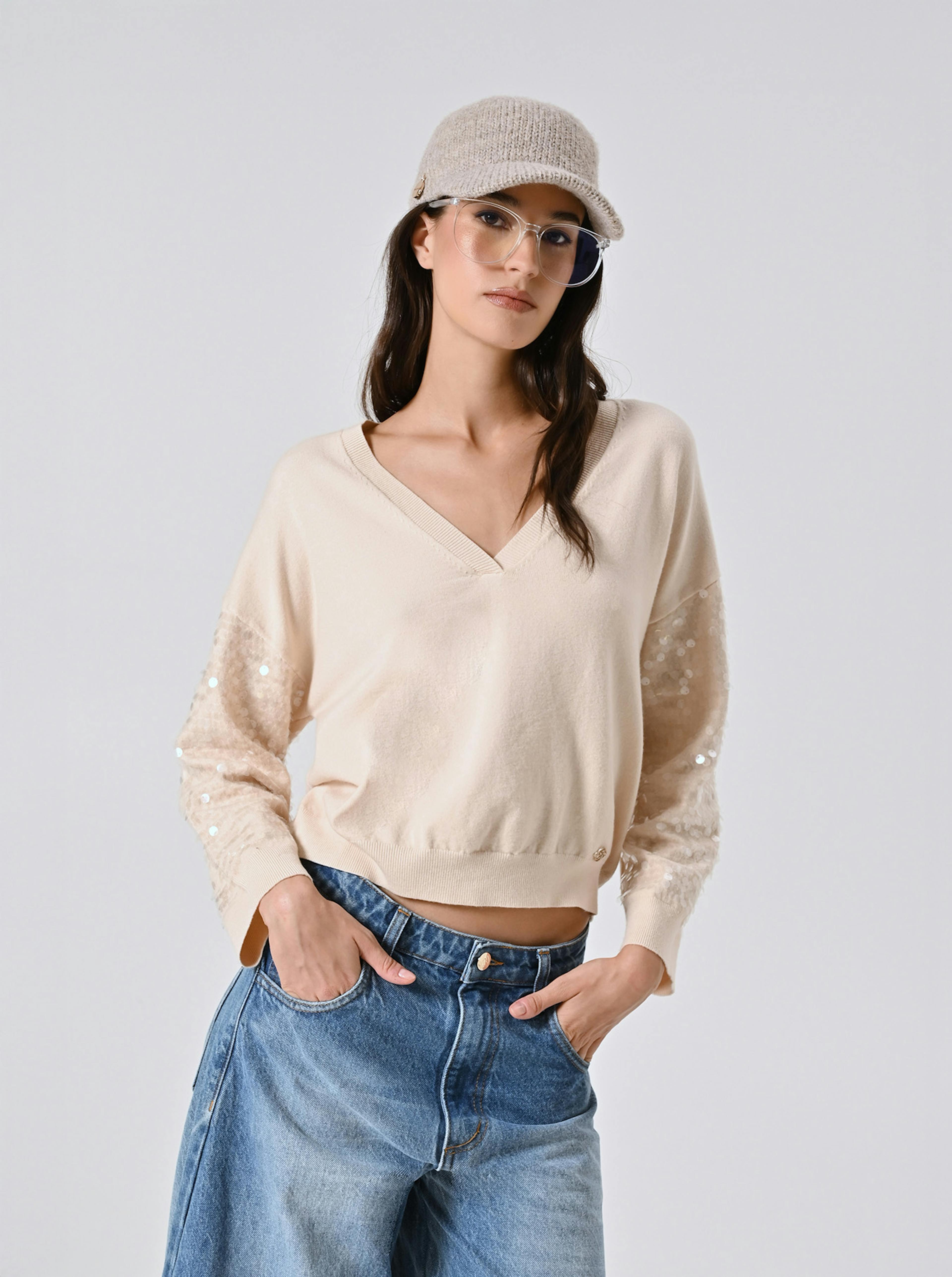 A woman in a beige sweater with sequin sleeves, blue jeans, and a knit cap poses confidently with hands in her pockets against a plain background.