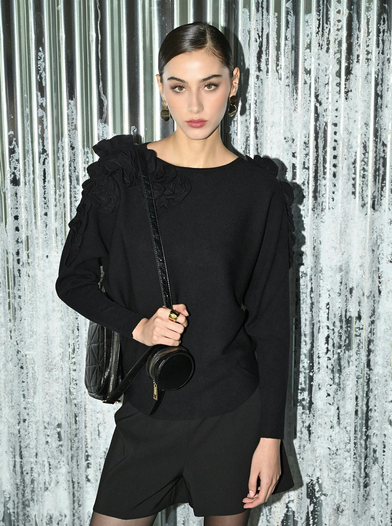 Woman in black sweater with ruffled shoulders and shorts, holding a black bag. She stands against a textured, metallic background, looking confident.