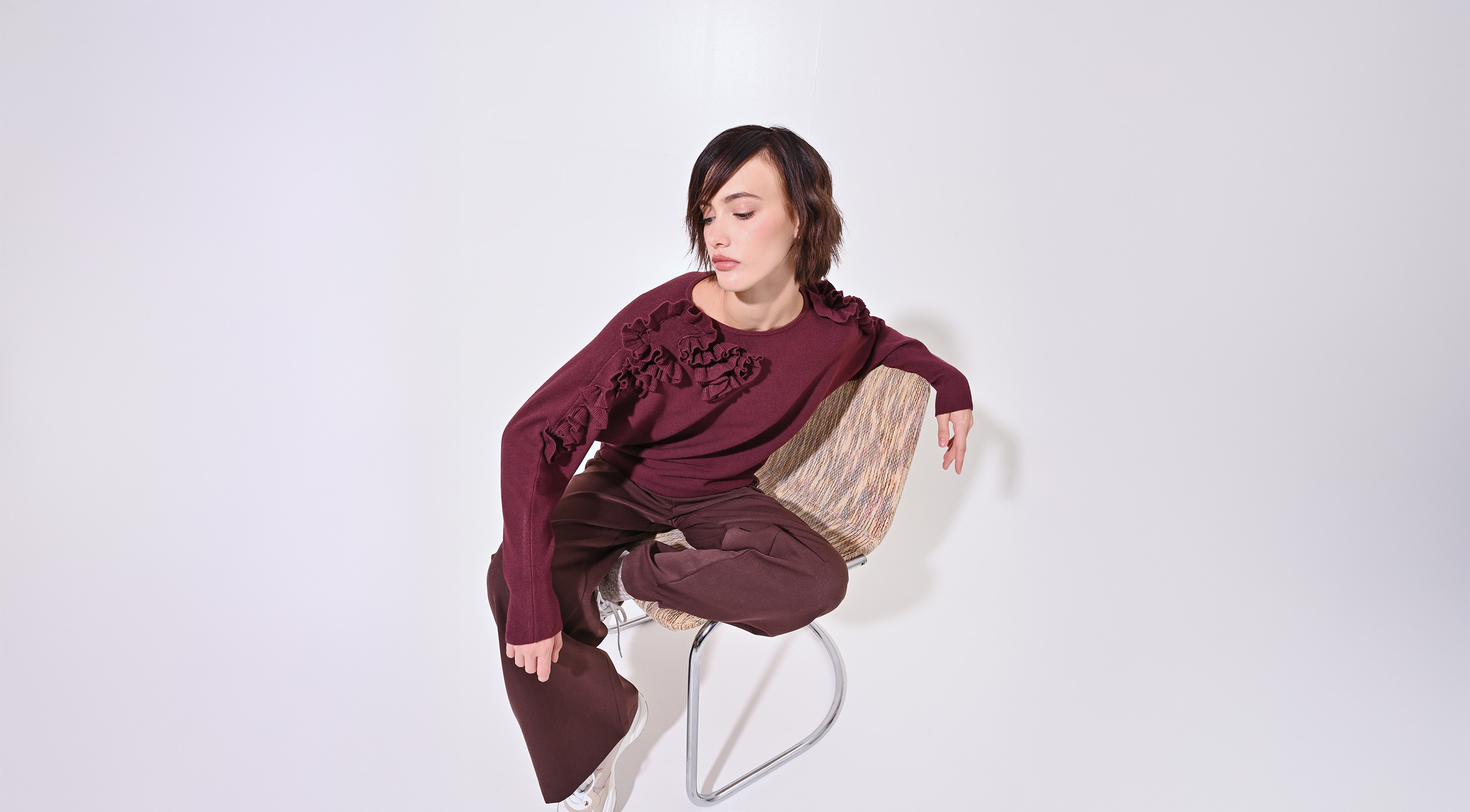 A person in a burgundy sweater with floral patterns sits on a modern chair against a white background, conveying a calm and contemplative mood.